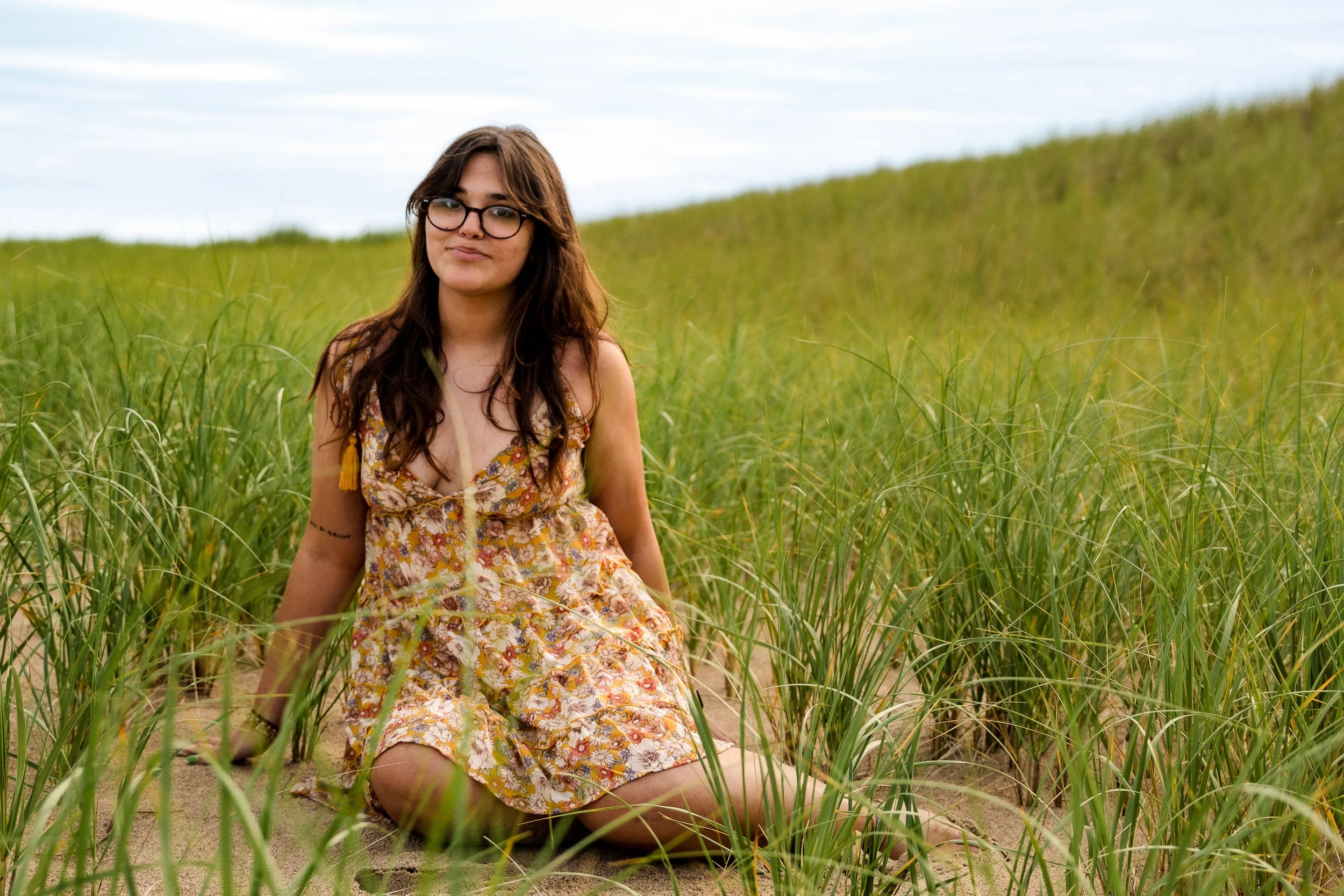 Young woman with long brown hair and glasses sitting in a grassy field, wearing a floral dress, with a calm expression on her face.