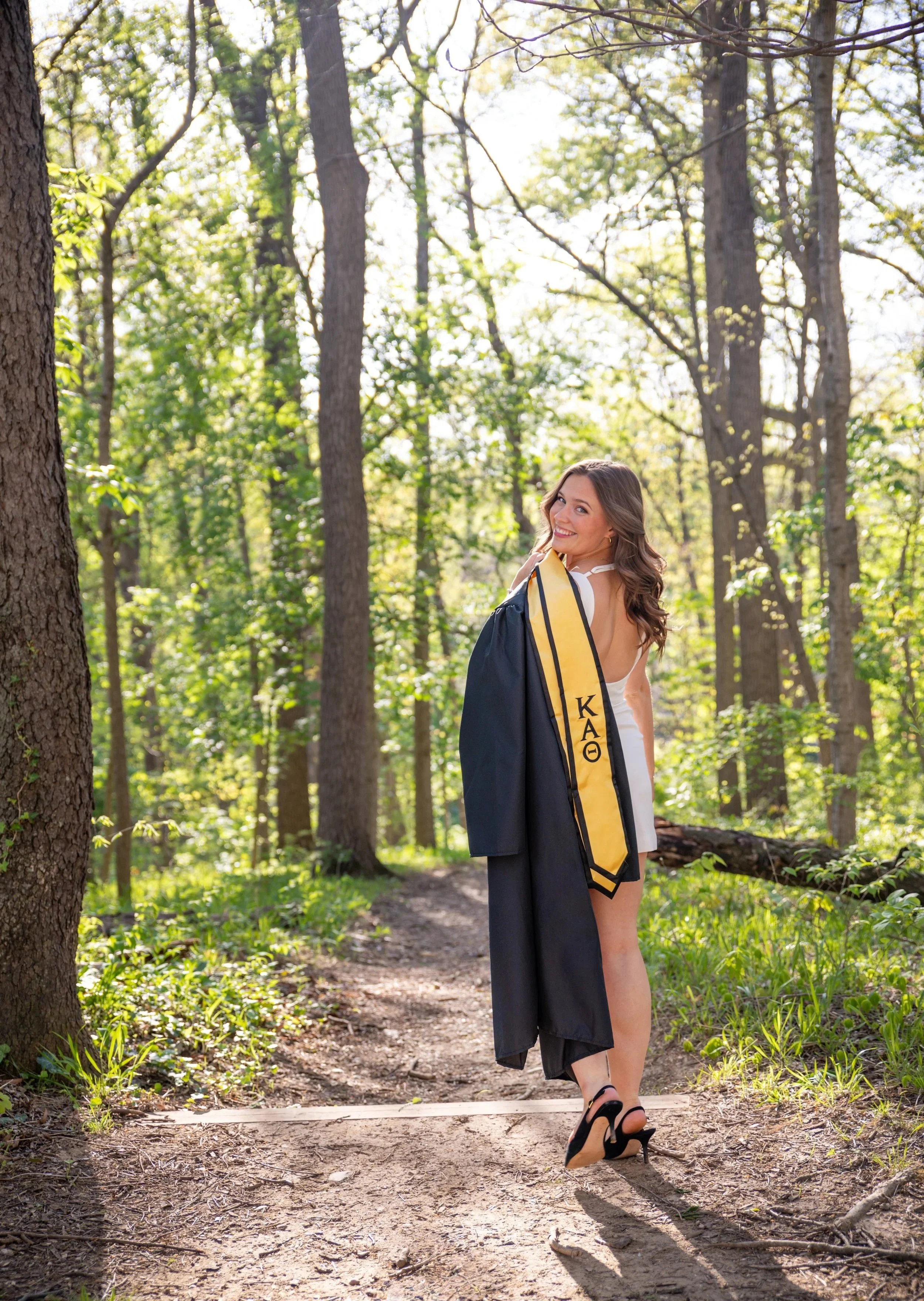 A young woman in white dress and high heels walks on a forest trail with a graduation gown draped over her shoulder and a yellow stole with Greek letters, surrounded by tall trees and bright green foliage.