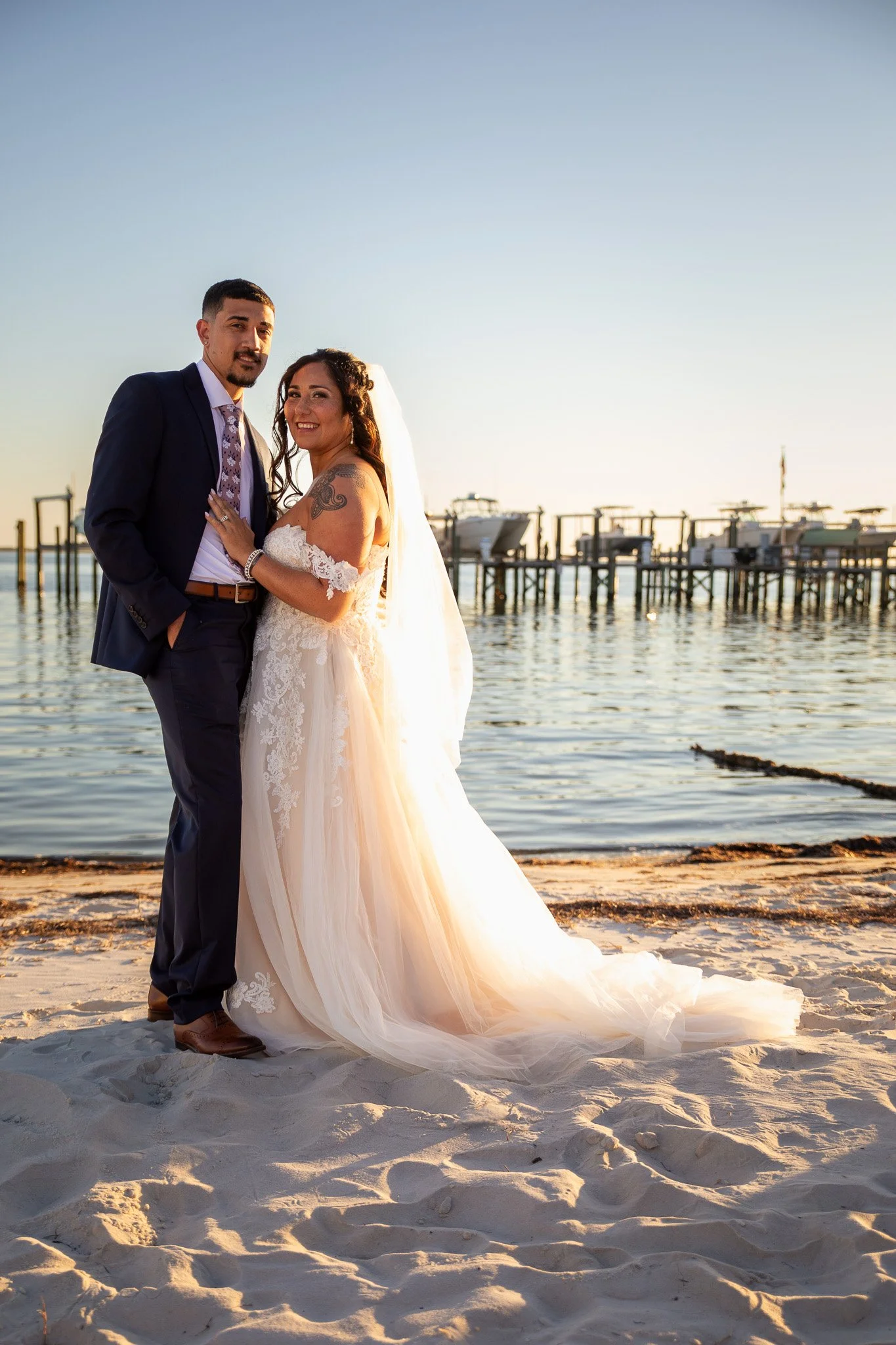 A newlywed couple standing on a sandy beach, with a pier and boats in the background during sunset, smiling and embracing each other.