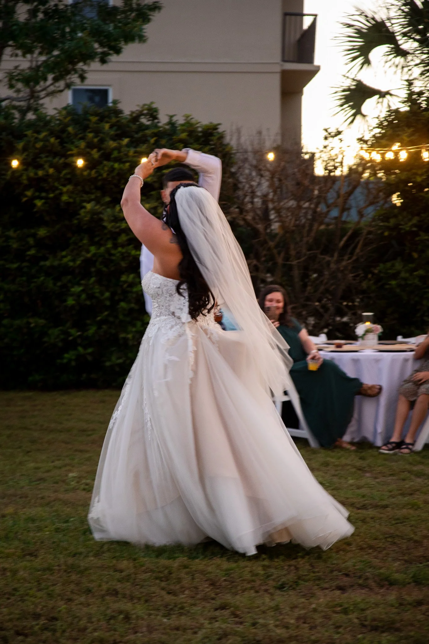 A bride dancing at an outdoor wedding reception during sunset, wearing a white strapless wedding gown with a long veil, with guests seated at tables in the background.