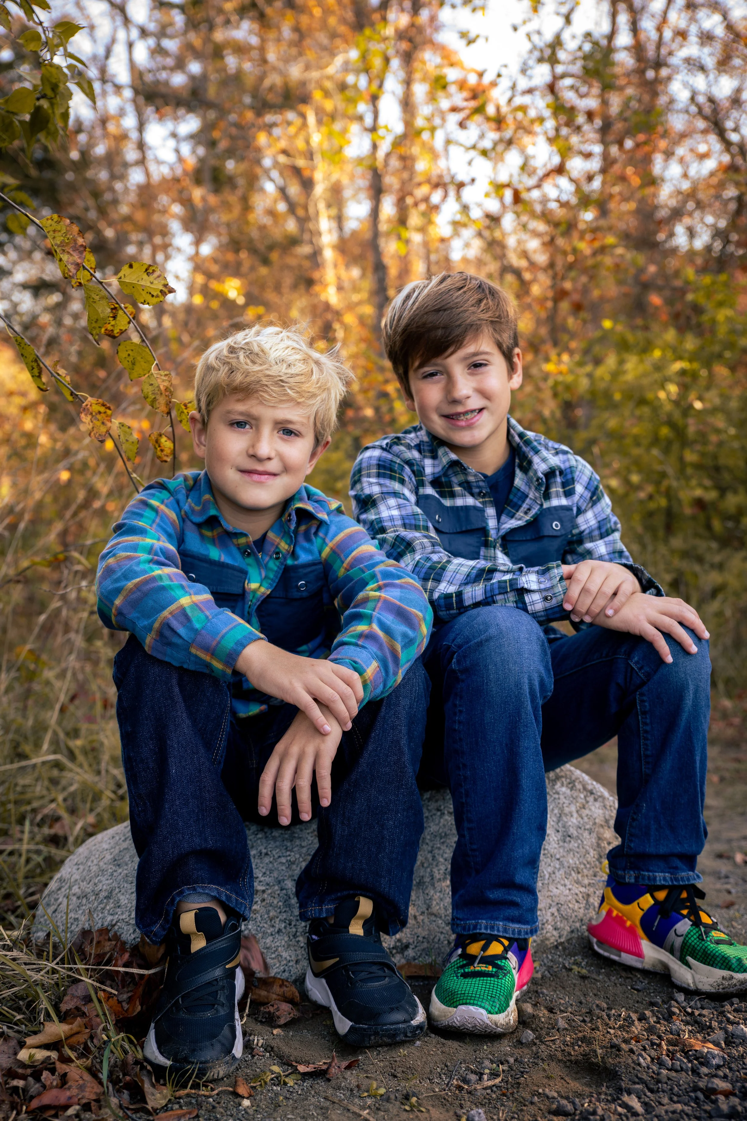 Two young boys sitting outdoors on a rock in a fall setting with colorful trees behind them, wearing plaid shirts and sneakers.