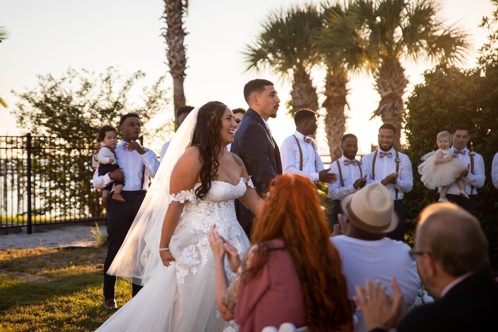 A bride and groom stand together during their outdoor wedding ceremony at sunset, surrounded by guests and palm trees.