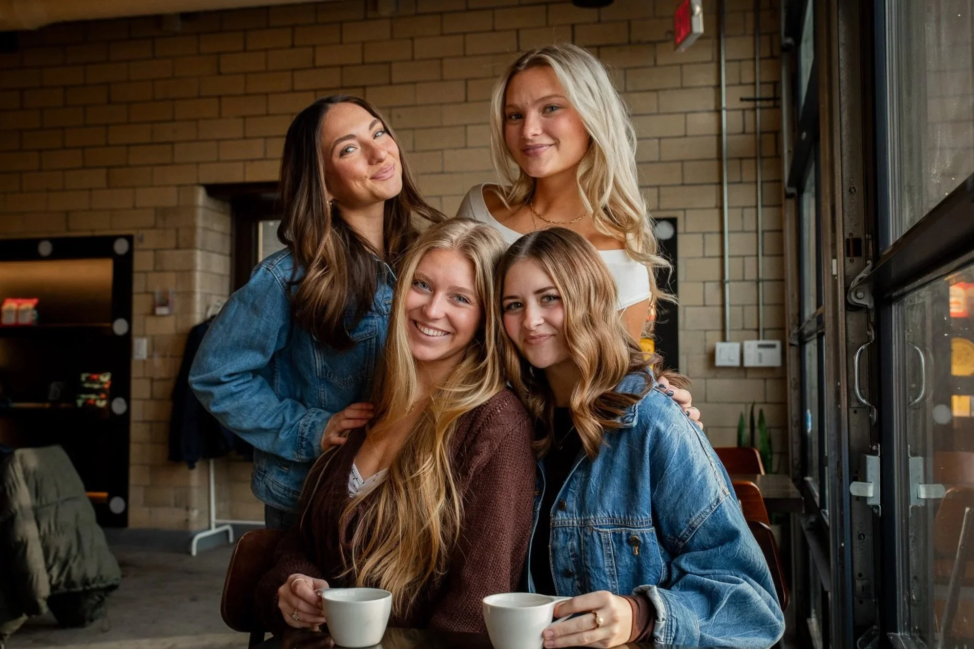 Five young women with blonde and brunette hair smiling and seated in a cafe, some holding cups of coffee, with a brick wall background and a vending machine on the side.