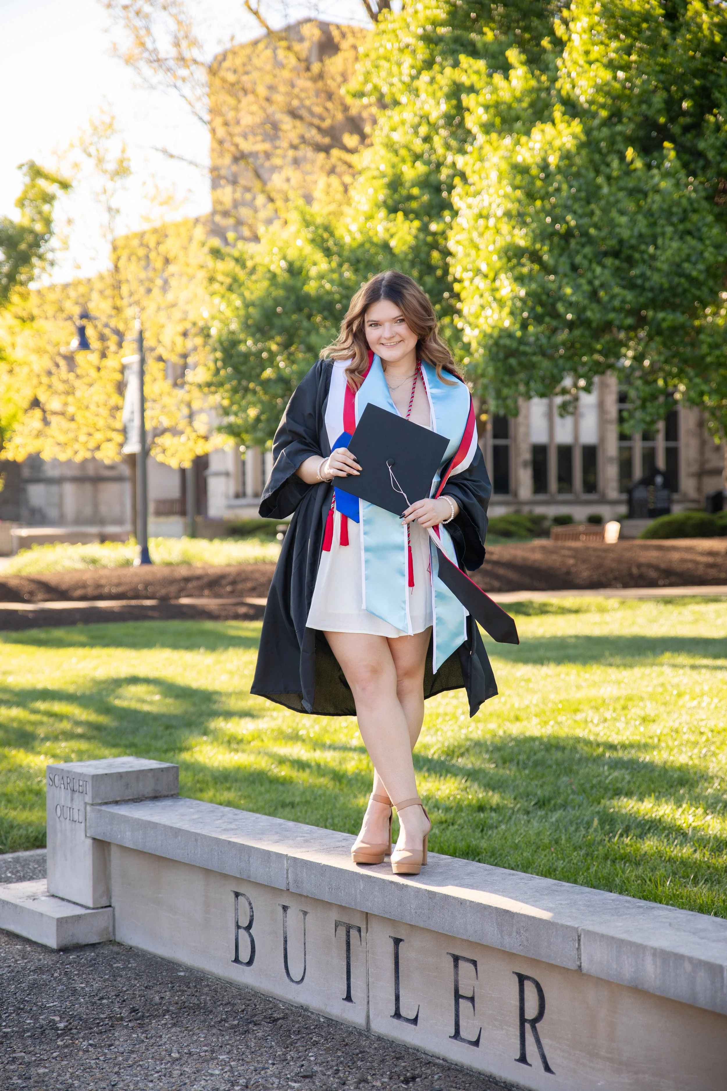 Young woman in graduation cap and gown standing on a low wall with a university building and green trees in the background. She is holding a diploma and smiling.
