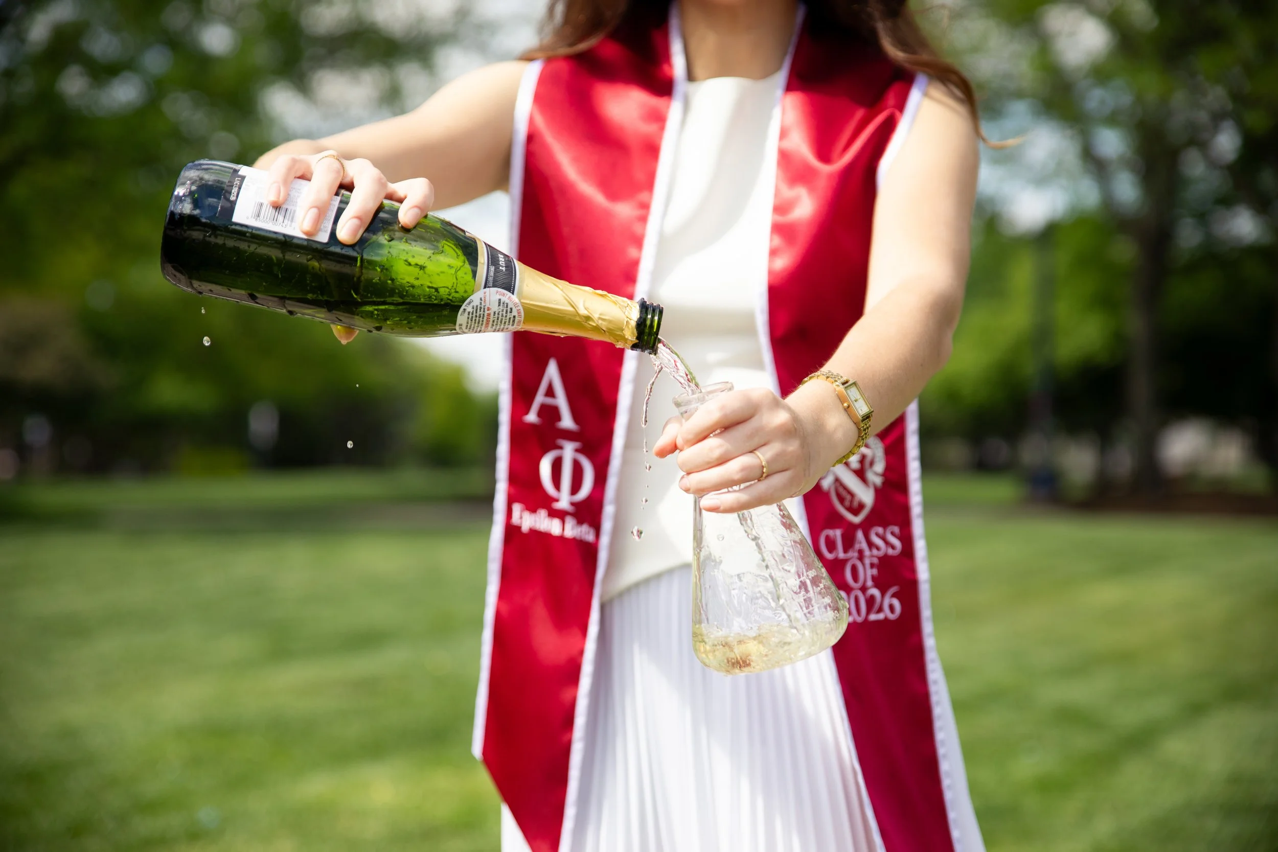 A woman wearing a red graduation stole with Greek letters pours champagne into a glass at an outdoor celebration.