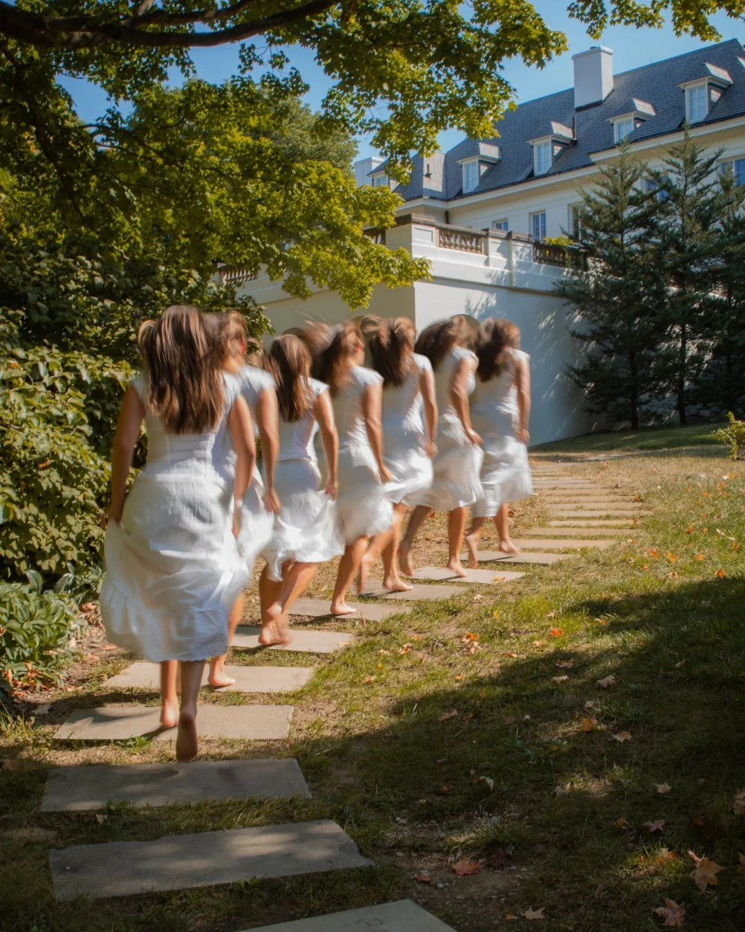 Multiple women dressed in white dresses are walking barefoot along a stone pathway in a garden, with a house and trees in the background on a sunny day.