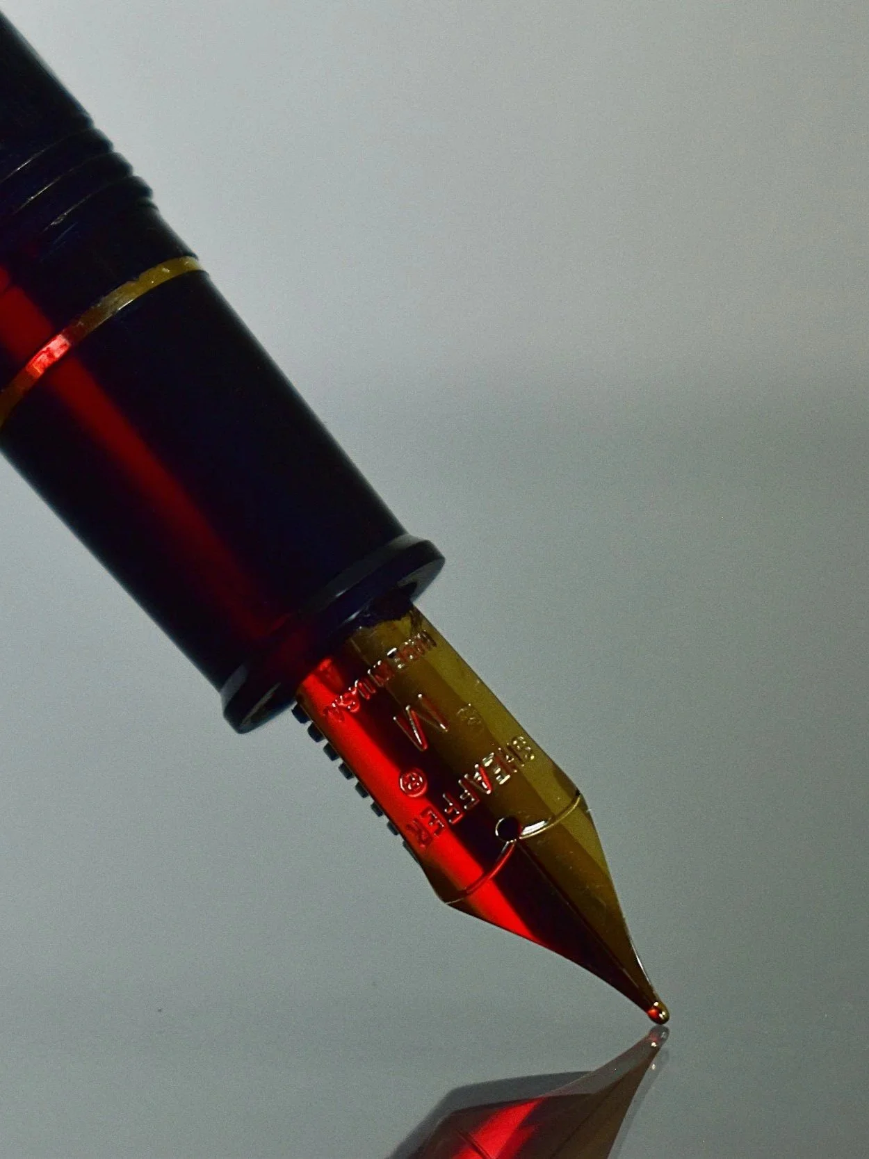 Close-up of a fountain pen nib, partially filled with ink, resting on a reflective surface with a plain background.