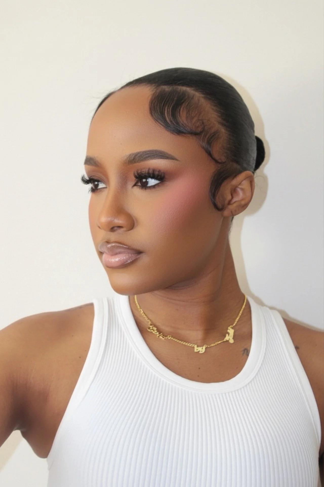 A woman with smooth skin, styled black hair with curls framing her face, wearing a white sleeveless top and gold jewelry, taking a selfie against a plain white background.