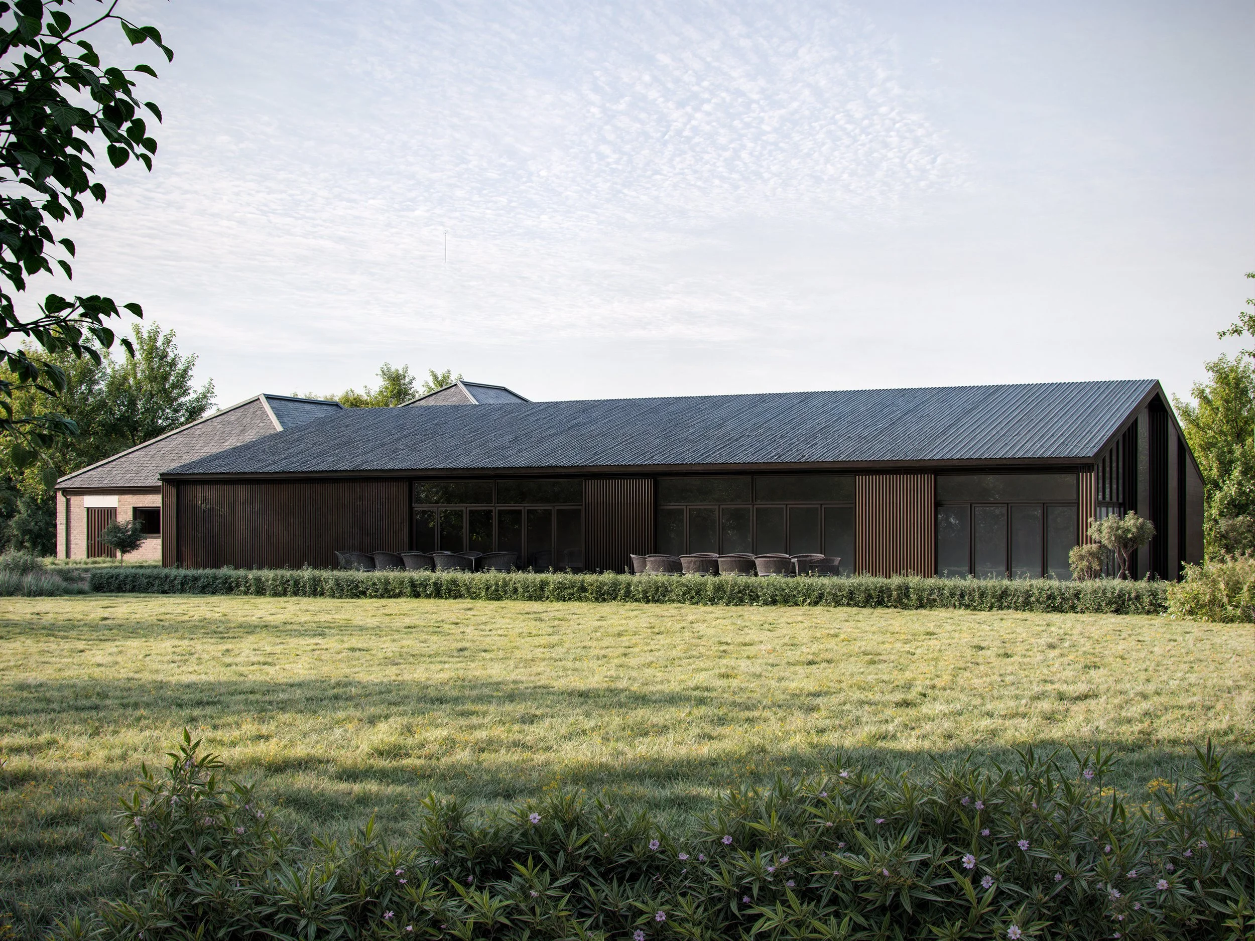 A modern house with a dark exterior and large glass windows is surrounded by a lush green lawn. There are trees and shrubs in the yard, and the sky above has scattered clouds.