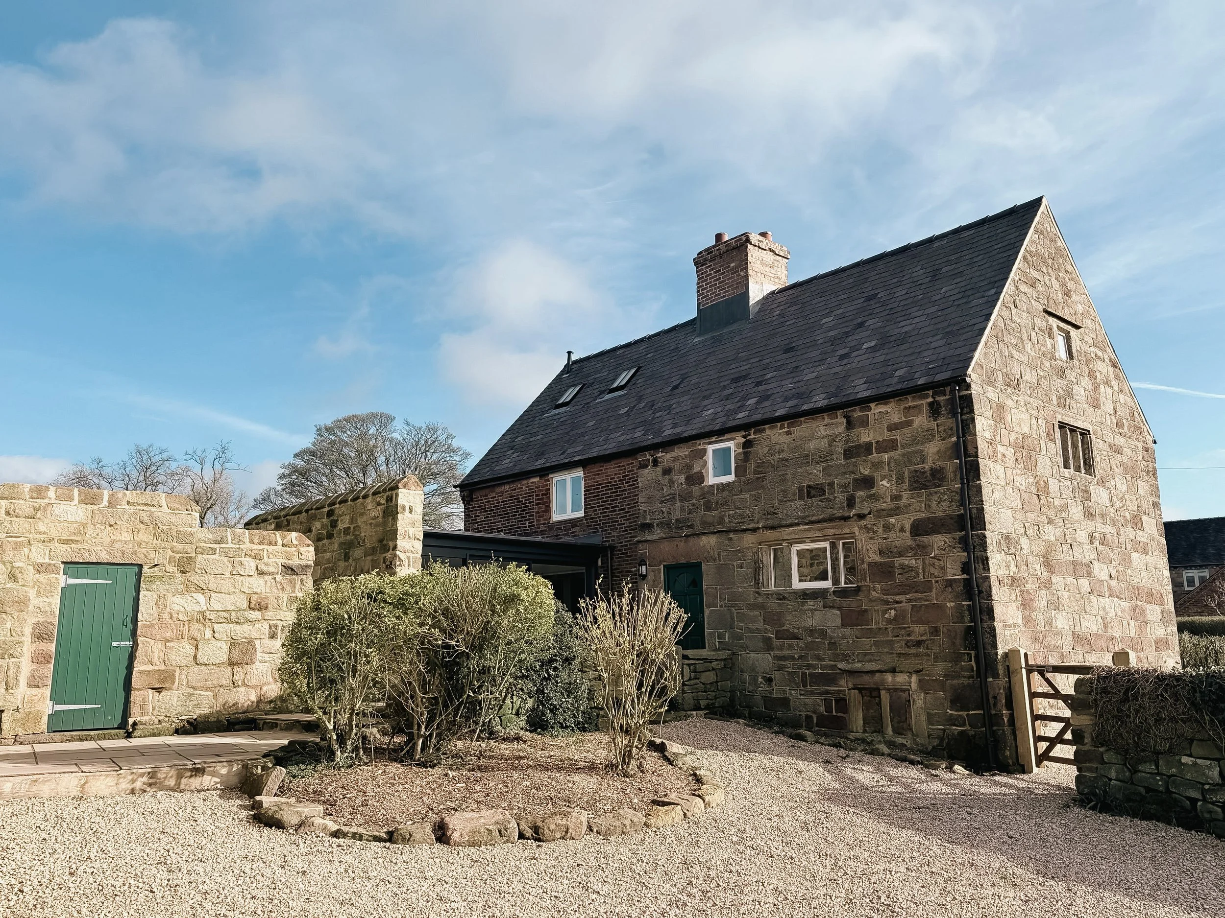 A stone house with a dark slate roof and a brick chimney under a blue sky with some clouds. There is a small green gate and some plants and a gravel yard in the foreground.