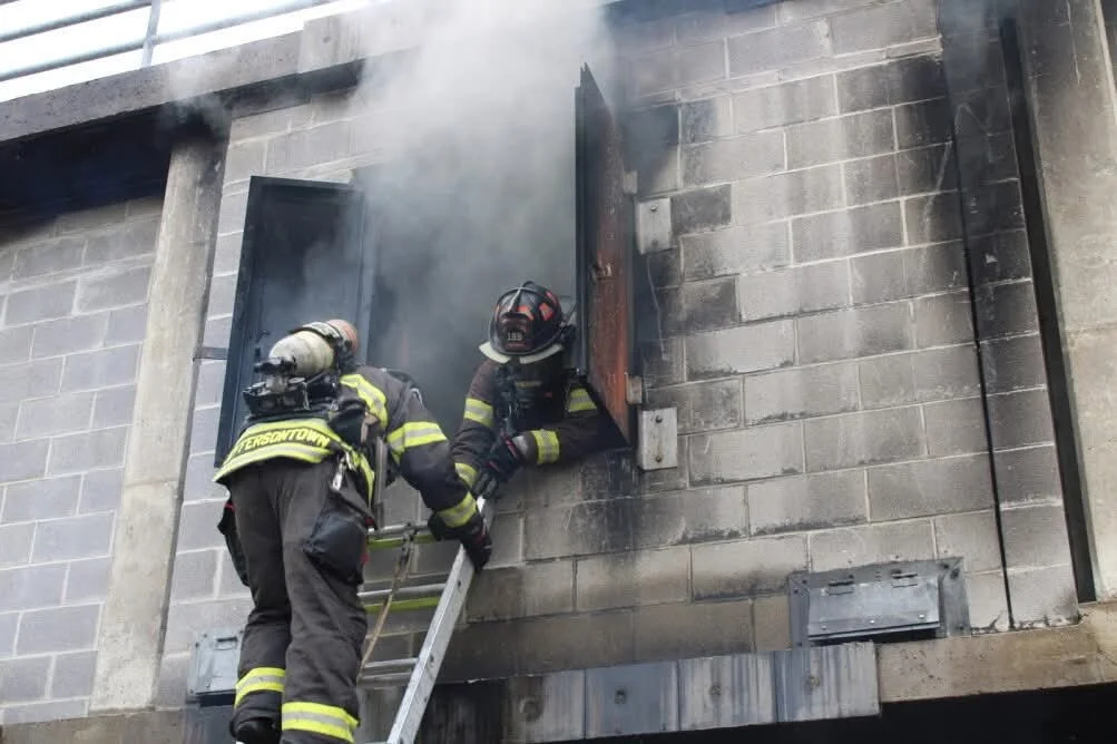 Two firefighters in black gear with yellow reflective stripes are using a small ladder to access a blackened window on a concrete wall with smoke coming out.