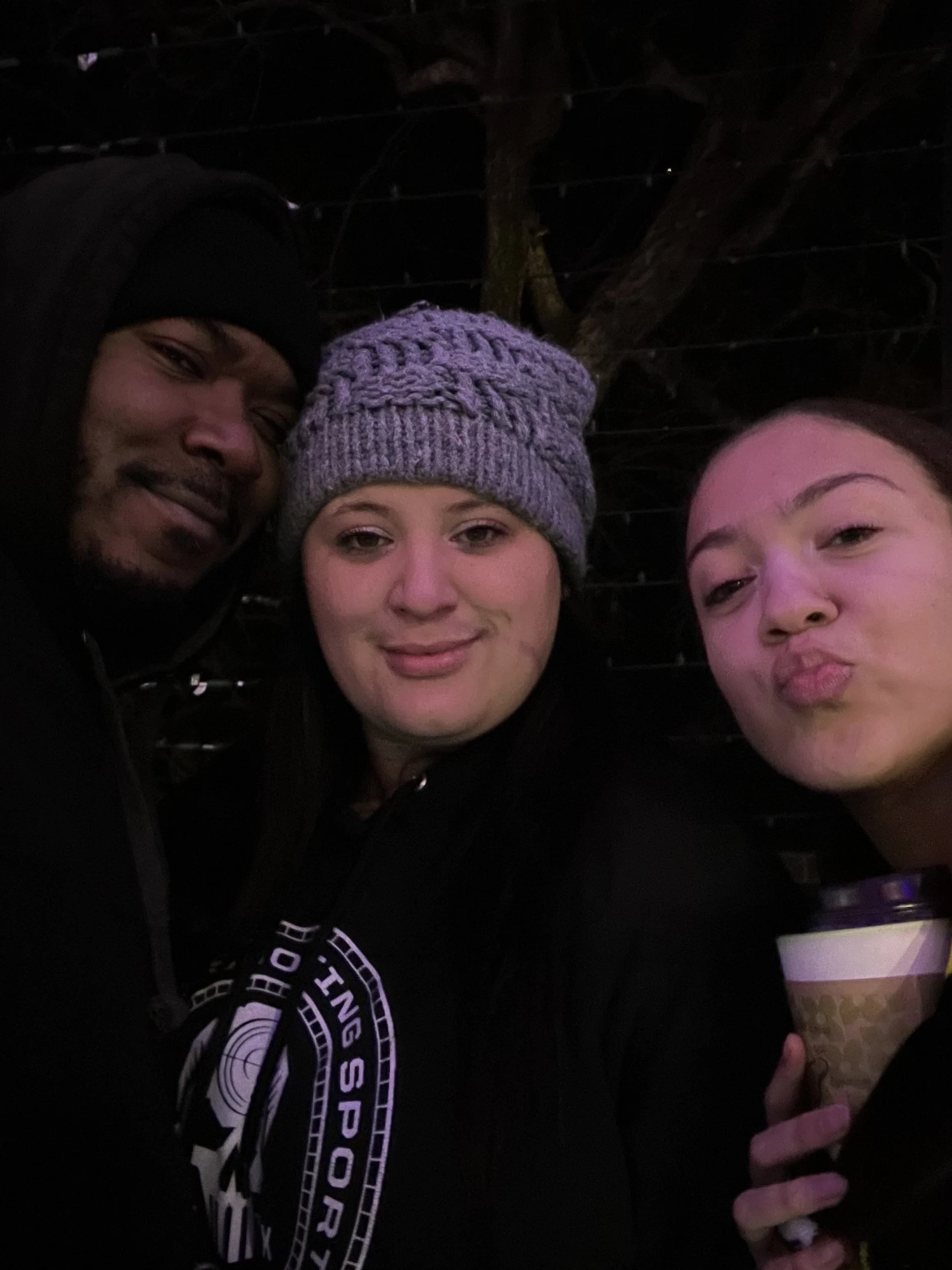 A group selfie of three people outdoors at night, with a dark background and a tree behind them. The person on the left wears a black jacket and black beanie, the person in the middle wears a purple knitted hat, and the person on the right has dark hair and is holding a cup.