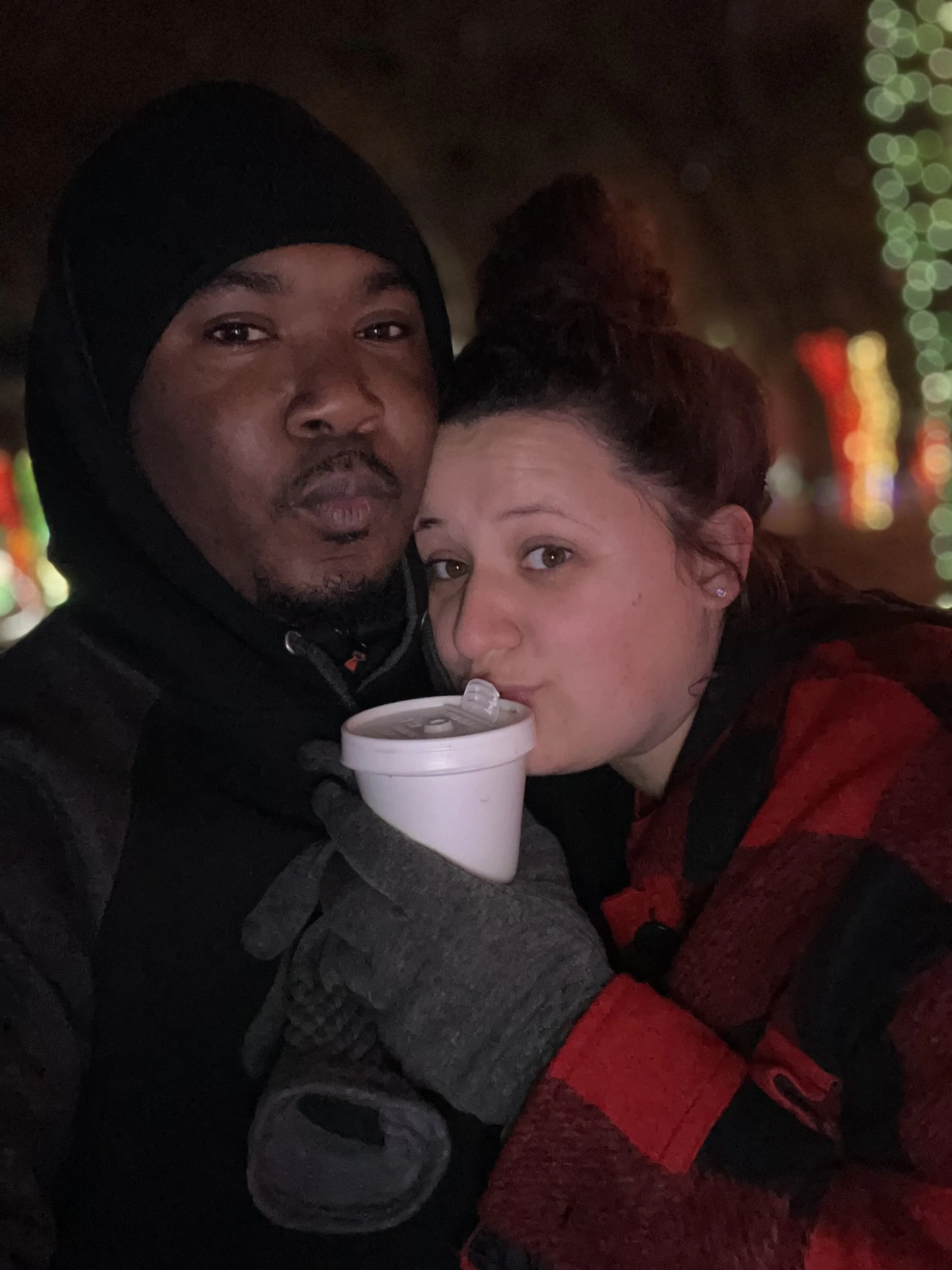 Two people, a man and a woman, taking a close-up selfie at night with colorful bokeh lights in the background. The man is wearing a black hoodie and a black beanie, and the woman is holding a white cup near her mouth, both looking into the camera.