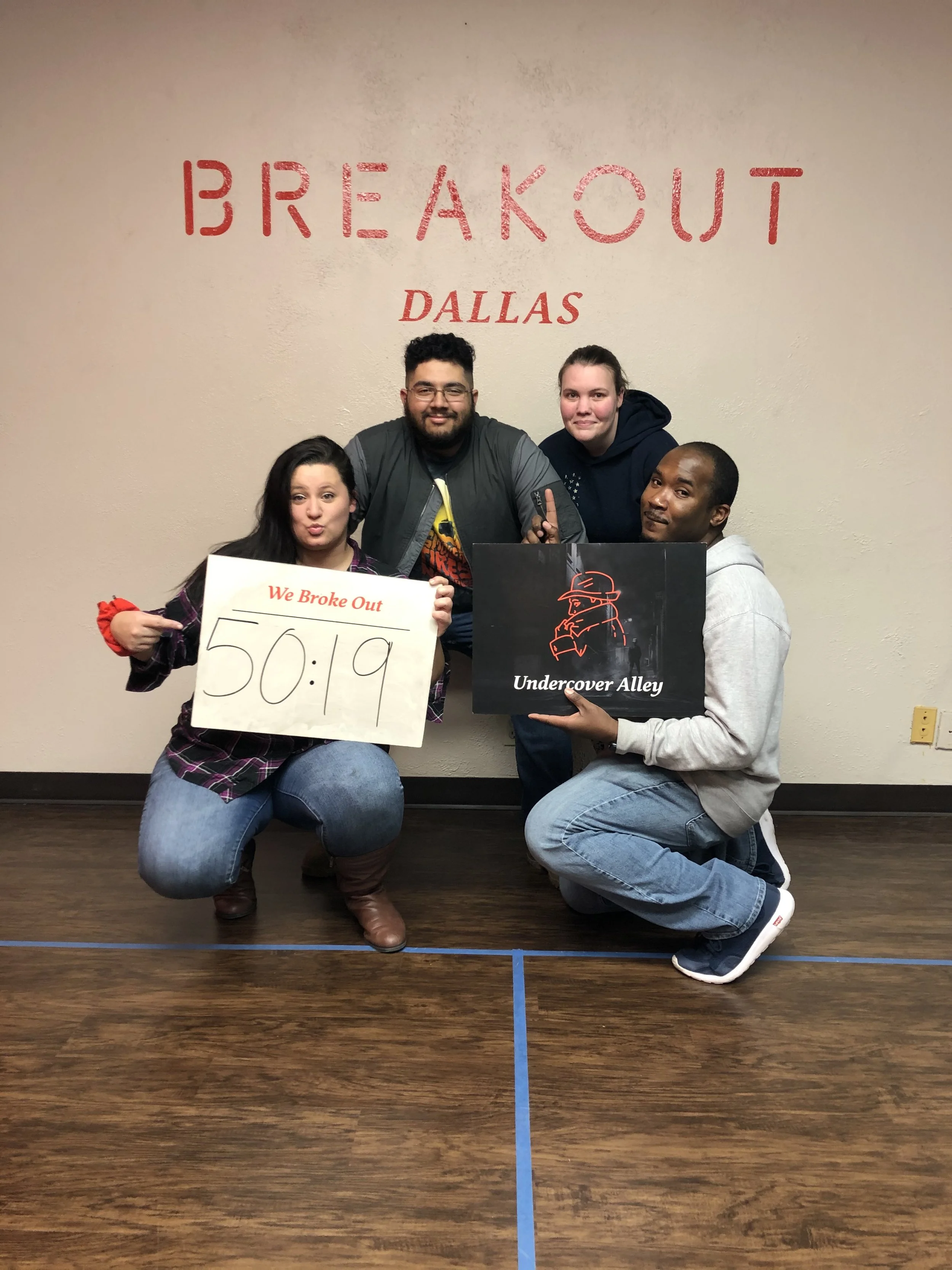 Group of four smiling people in front of a wall with red text reading 'BREAKOUT DALLAS'. One person holds a sign with 'We Broke Out 50:19' written on it, and another holds a black and red poster with a red outline of a person in a hat and the text 'Undercover Alley'. They are kneeling and standing on a wooden floor with blue tape lines.