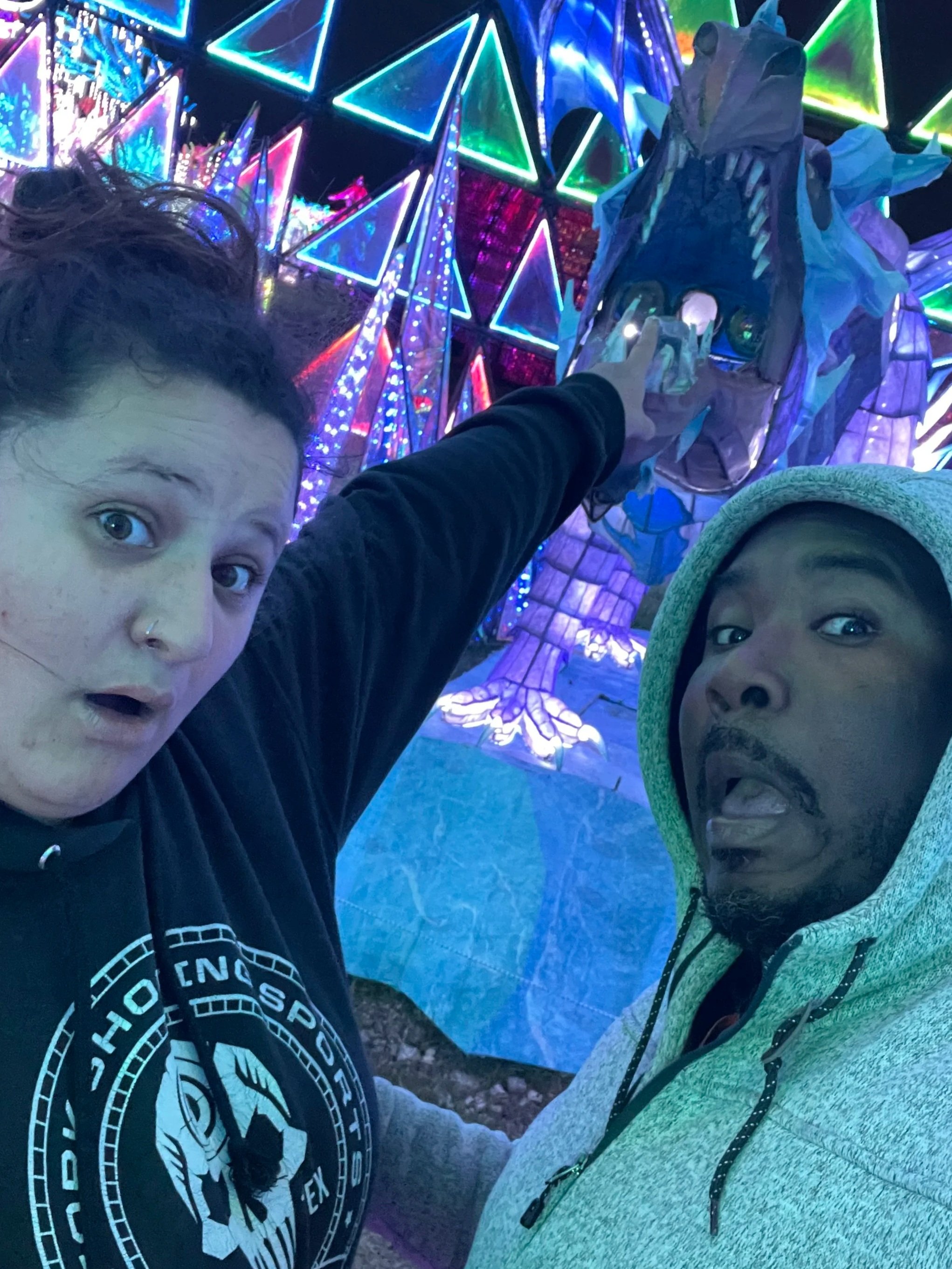 Two people taking a selfie in front of a colorful, neon-lit art installation featuring a dinosaur skeleton.