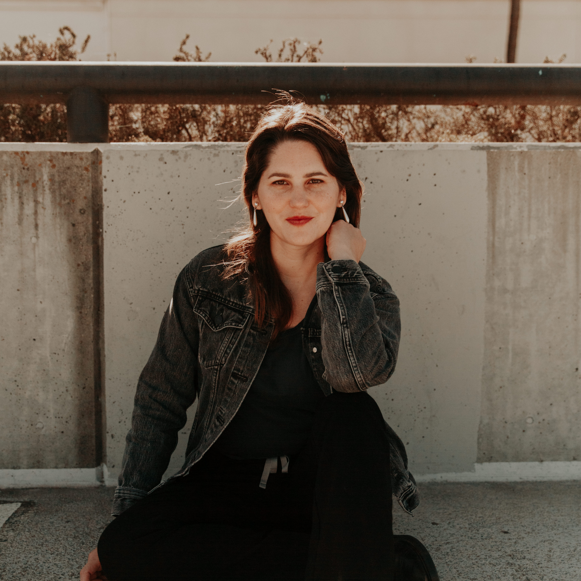 A woman with dark hair and light skin, wearing earrings and a black jacket, sitting outdoors near a concrete wall, with trees and a fence in the background.