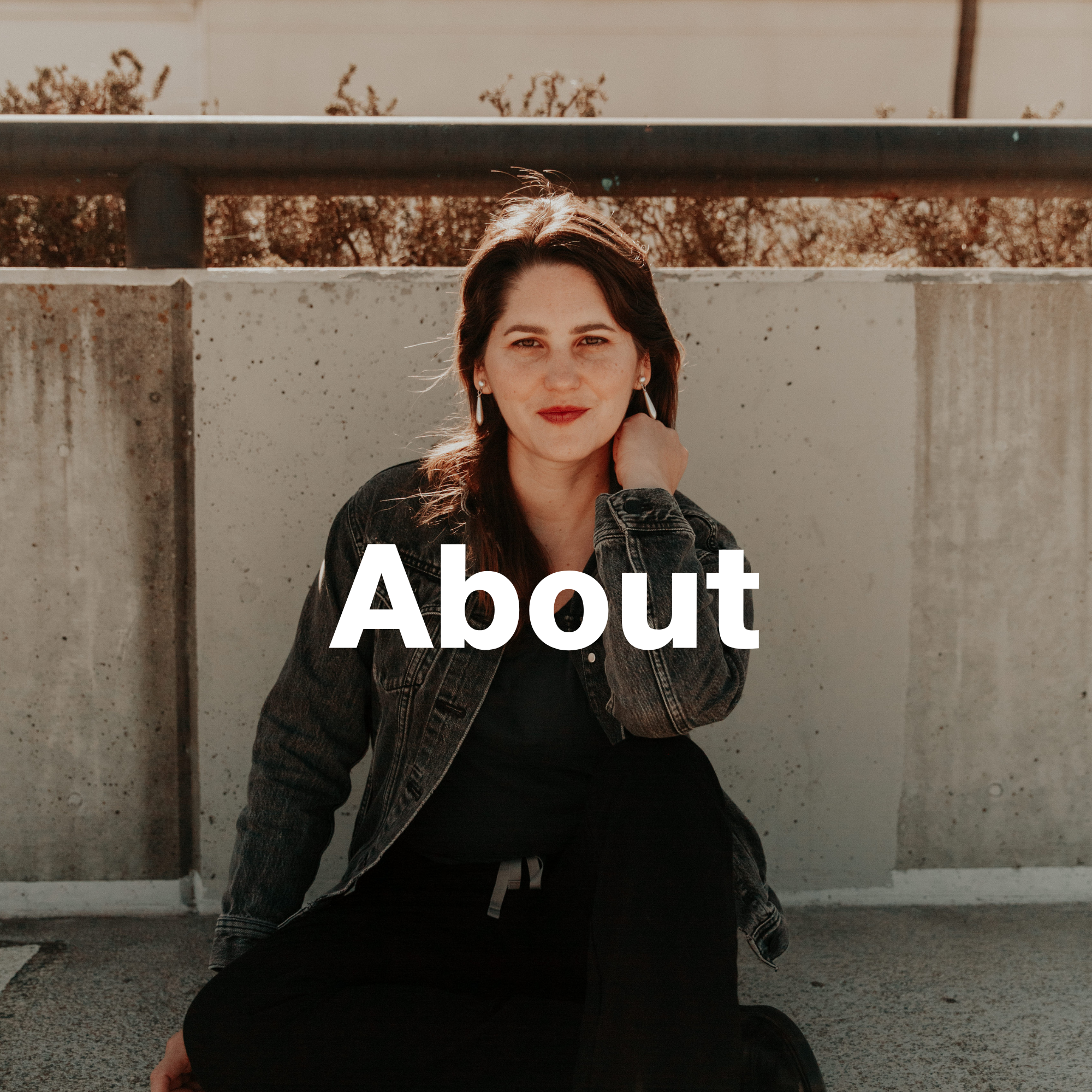 A woman with dark hair and red lipstick, wearing earrings and a denim jacket, sitting outdoors against a concrete wall with a metal railing above, in warm sunlight.