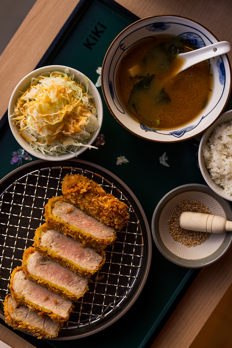 Japanese meal featuring sliced fried pork cutlet on a wire rack, a bowl of miso soup with seaweed and a spoon, a bowl of steamed white rice, a small dish of sesame seeds and a pestle, and a bowl of shredded cabbage salad topped with shredded cheese and dressing.