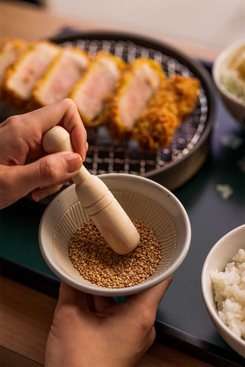 Person using a wooden pestle to grind sesame seeds in a small bowl, with fried breaded meat cutlets and bowls of rice and cabbage salad in the background.