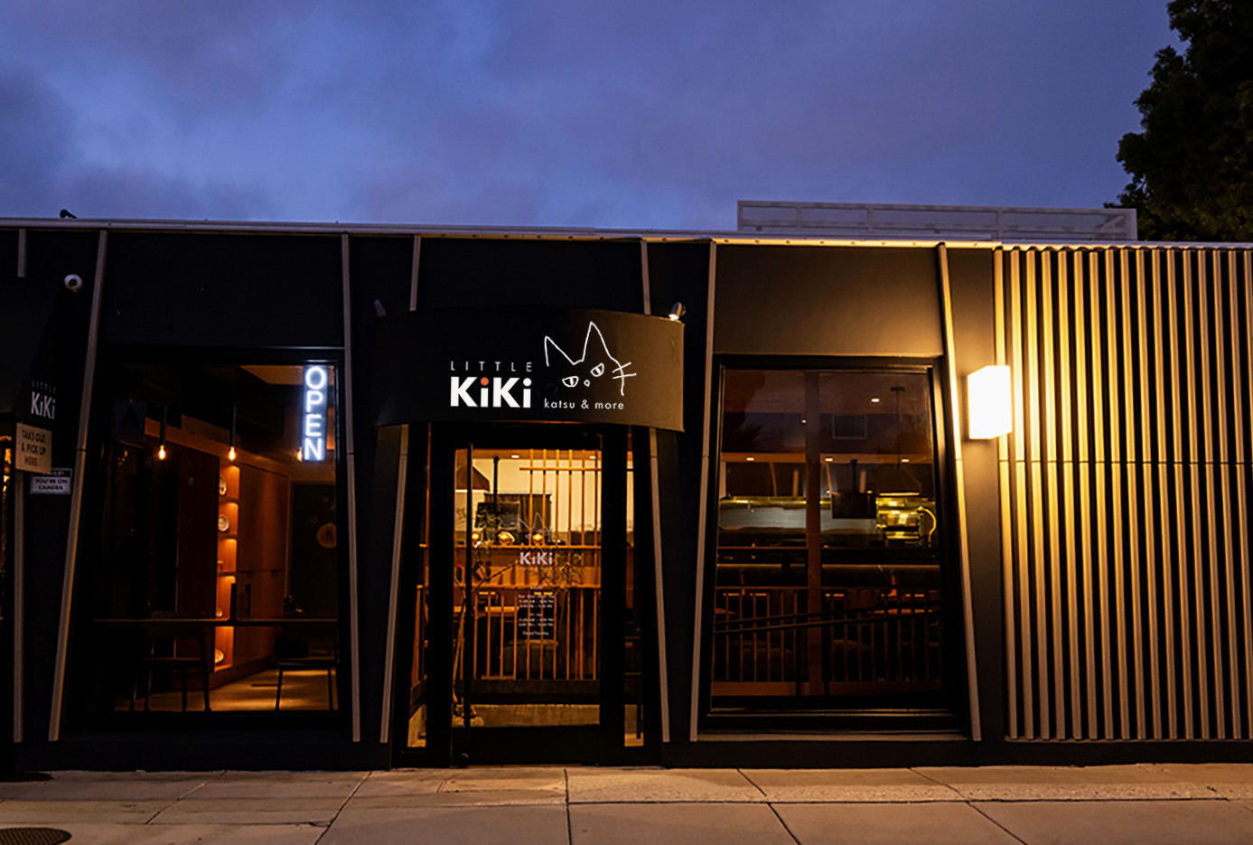 Exterior of a modern restaurant with a black facade, illuminated sign reading 'LITTLE KiKi katsu & more', and large glass windows showing the interior.