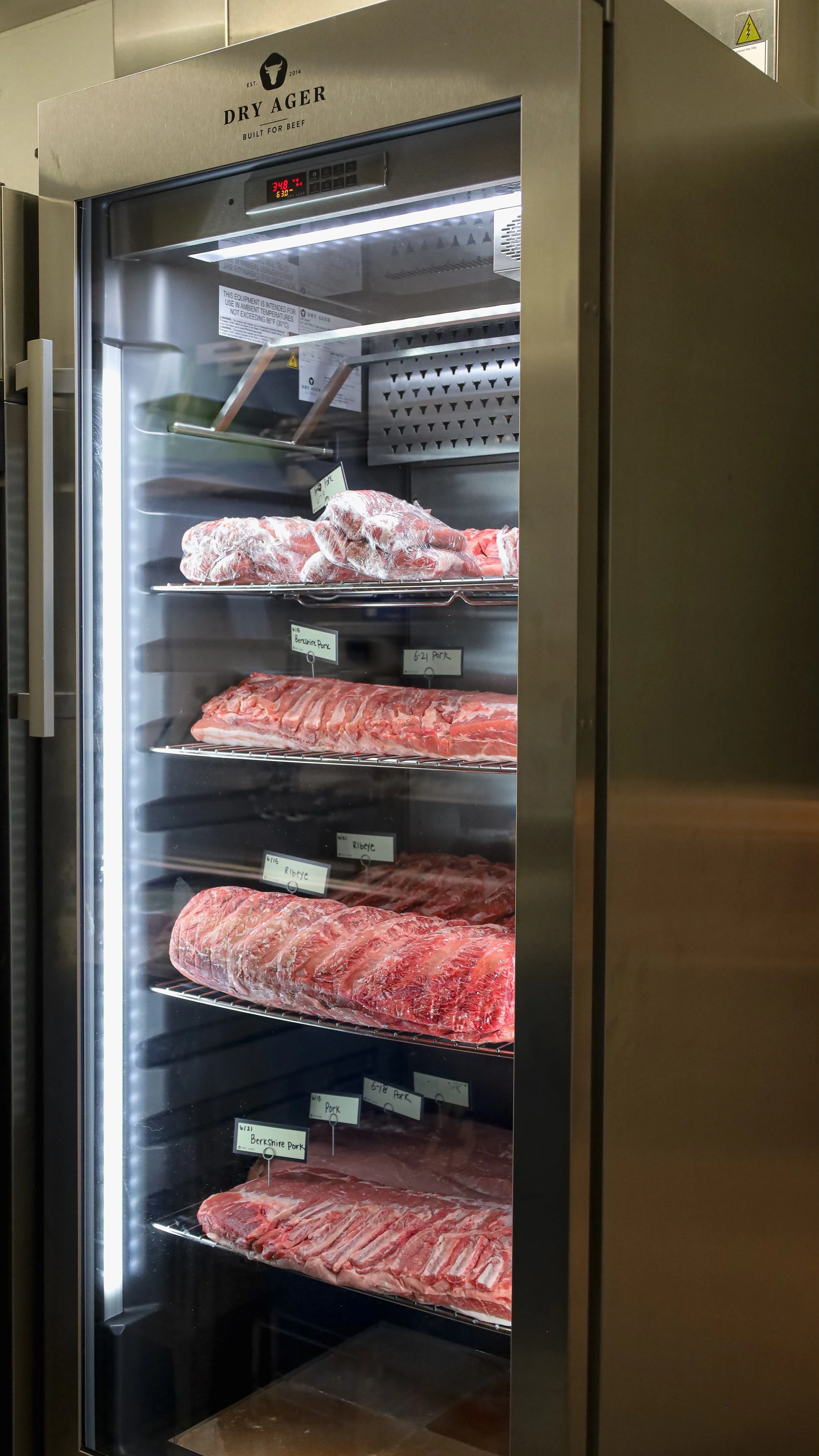 A commercial meat fridge with multiple shelves stocked with various cuts of beef and pork, labeled with tags indicating different types of meat.