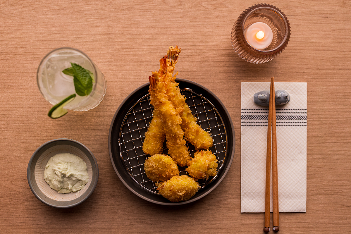 A plate of tempura shrimp and vegetables with dipping sauce, a glass of lime and mint drink, a candle in a glass holder, and a pair of chopsticks on a paper napkin on a wooden table.
