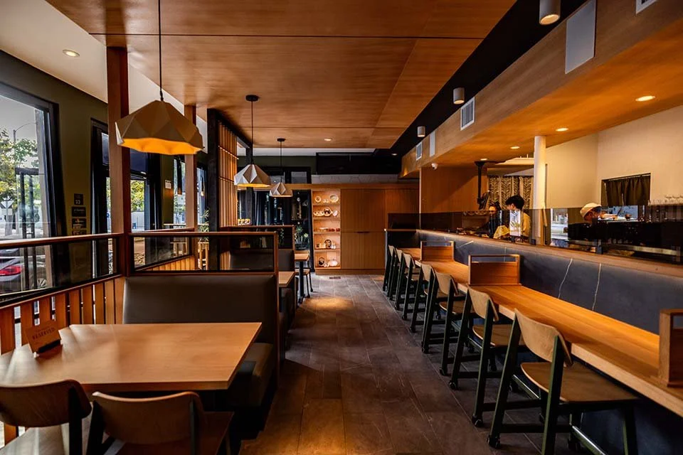 Inside a modern cafe with a wooden ceiling and walls, large windows, a bar with stools, and a seating area with tables and chairs.