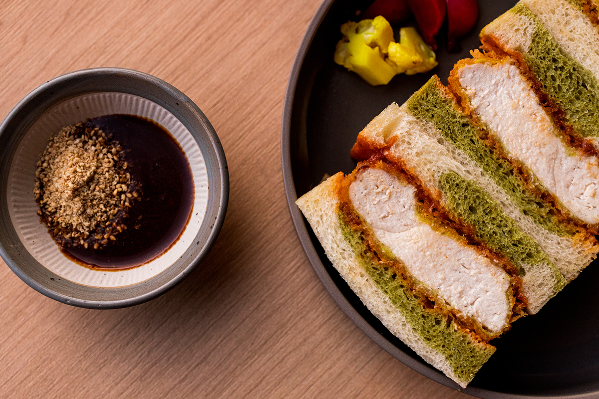 Three layers of green and white sandwich, with fried chicken inside, on a black plate with pickles and tomatoes. A small bowl of dipping sauce with crushed peanuts is next to it on a wooden surface.