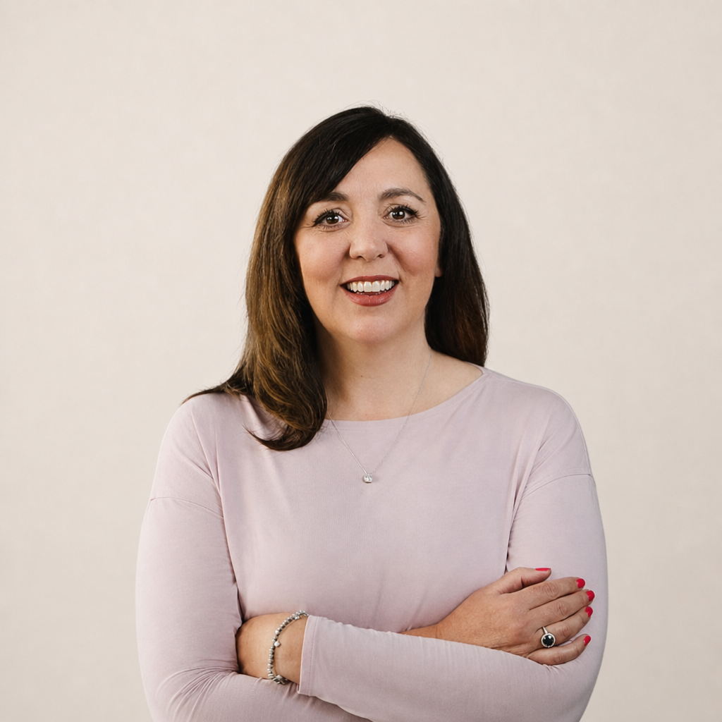 Portrait of a woman with dark brown hair, wearing a pale pink top, smiling with arms crossed against a plain light background.
