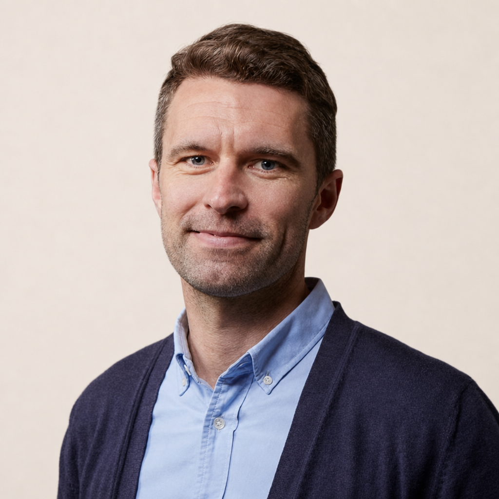 A middle-aged man with short brown hair, blue eyes, and light stubble, wearing a light blue button-up shirt and a dark navy cardigan, smiling slightly against a plain light background.