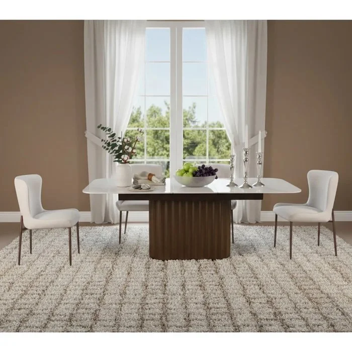Sala de comedor con mesa blanca y dos sillas blancas, si hay un jarrón con follaje, una bowl con uvas verdes y moradas, y candelabros plateados, con ventana grande y cortinas blancas.