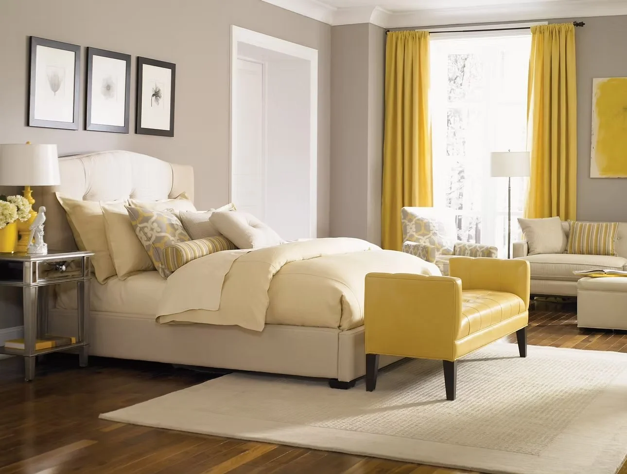 Habitación con paredes grises, cama con cabecera blanca y ropa de cama en tonos beige y gris, cortinas amarillas, sillón y banco en tonos amarillos, mesa de noche con lámpara y cuadros con insectos en la pared.