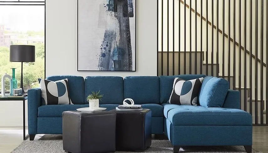 Sala moderna con sofá azul, cojines decorativos en blanco, negro y azul, mesas cuadradas negras, un libro y teléfono en ellas, ventana grande con vista a áreas verdes, cuadro abstracto en la pared, escaleras de madera y barandilla de barras negras.