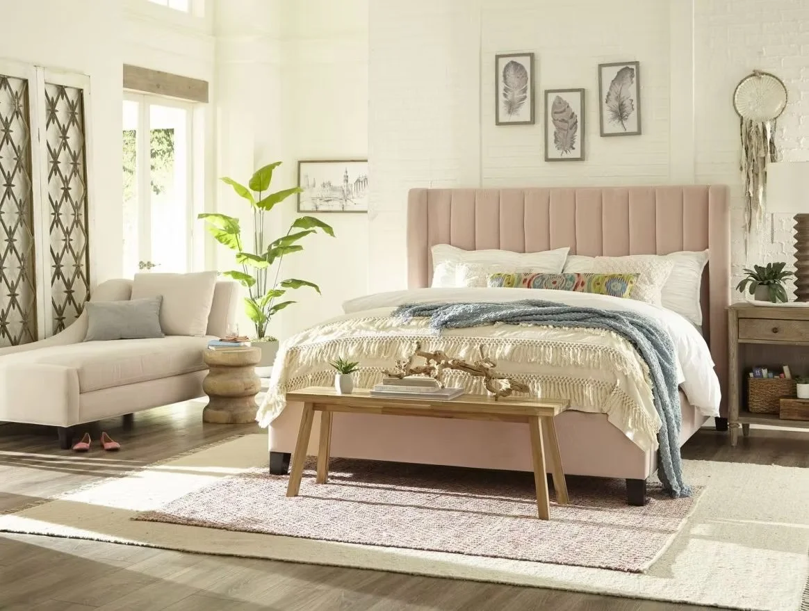 Habitación con cama con cabecera rosa, pared decorada con cuadros de hojas, sofá blanco, planta grande, mesita con libros y decoración, alfombra, y ventanas con cortinas.