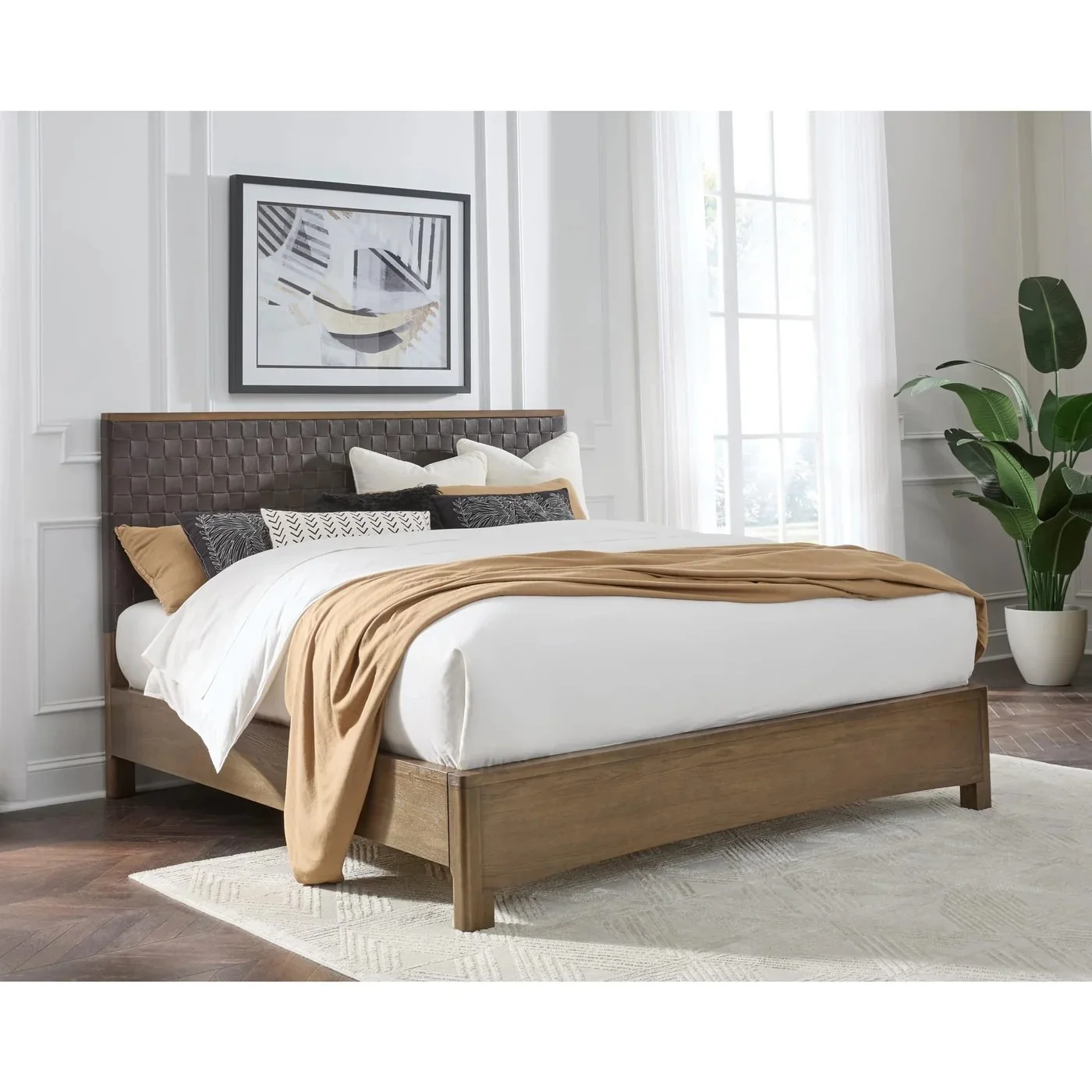 Dormitorio con cama grande, cabecera de cuero acolchonado, varias almohadas, colcha beige, cortinas blancas y un cuadro moderno en la pared. Planta grande en una maceta blanca junto a la ventana.