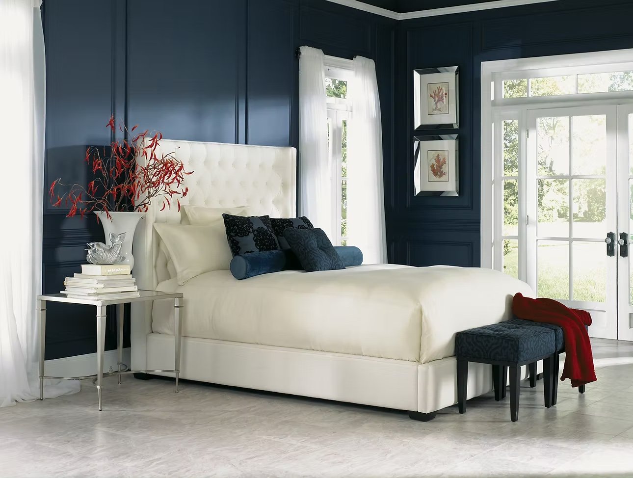 Dormitorio con paredes azul marino, cama blanca con cabecera acolchada, cortinas blancas, decoraciones de marcos y cuadros con motivos de hojas, ventana grande con vista al exterior, mesa de noche con libros y decoración de ave, cojines oscuros, banco a los pies con cobija roja.