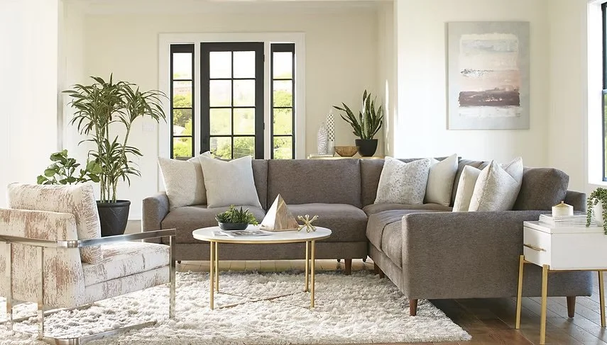 Sala de estar con sillones grises, madera clara, y plantas decorativas junto a ventanas grandes con vista al exterior.