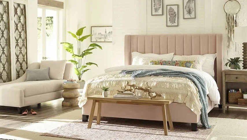 Dormitorio con cama de cabecera acolchada en color rosa, paredes blancas, paredes decoradas con cuadros de hojas, una planta grande junto a la ventana, un sillón blanco con cojines, mesa de noche de madera, y un mueble de madera con decoración y libros.