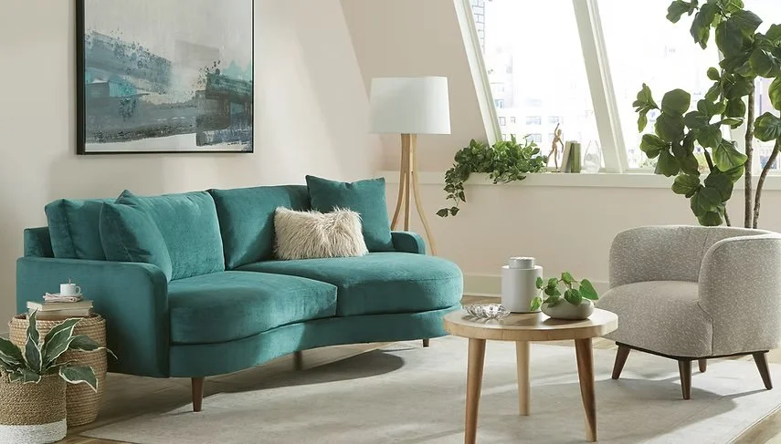 Sala moderna con sillón de color azul turquesa, dos sillones grises, mesa de centro de madera con plantas y objetos decorativos, ventanas grandes con vista a edificios urbanos, y árbol de interior en la esquina derecha.