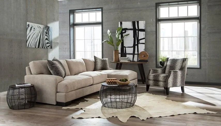 Sala moderna con sofá beige, mesa de centro de metal, dos sillones con estampado, arte abstracto en la pared, plantas y ventanas grandes con vista a edificios.