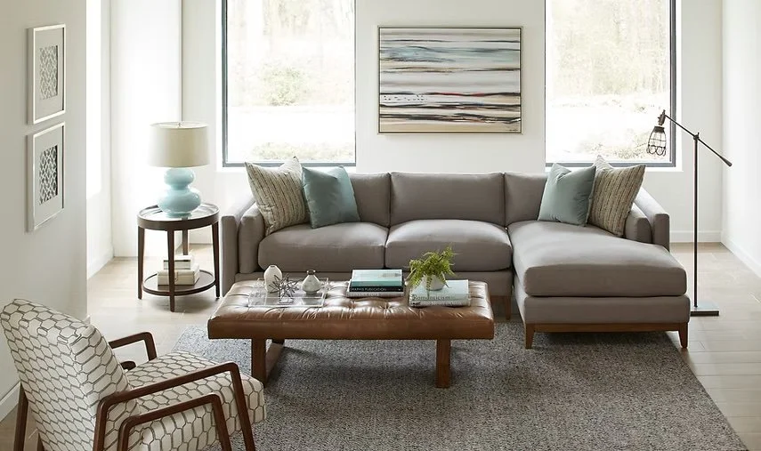 Sala de estar con sofá gris, mesa de centro de madera, sillón con patrón hexagonal, lámpara de pie, mesa auxiliar con lámpara, cuadros en la pared, y cojines de colores pastel.
