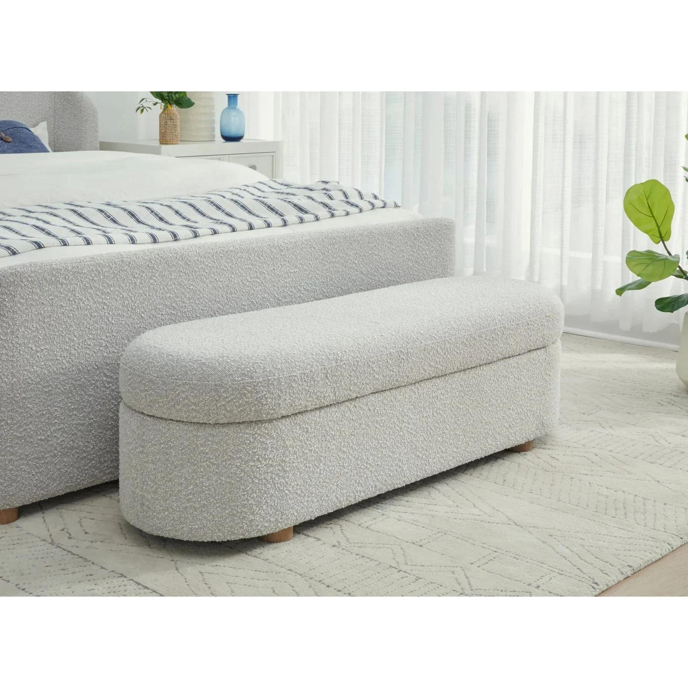 Sala de estar con sofá, cojín, mueble de madera blanca, plantas verdes, cortinas blancas transparentes y alfombra beige con patrón sutil.