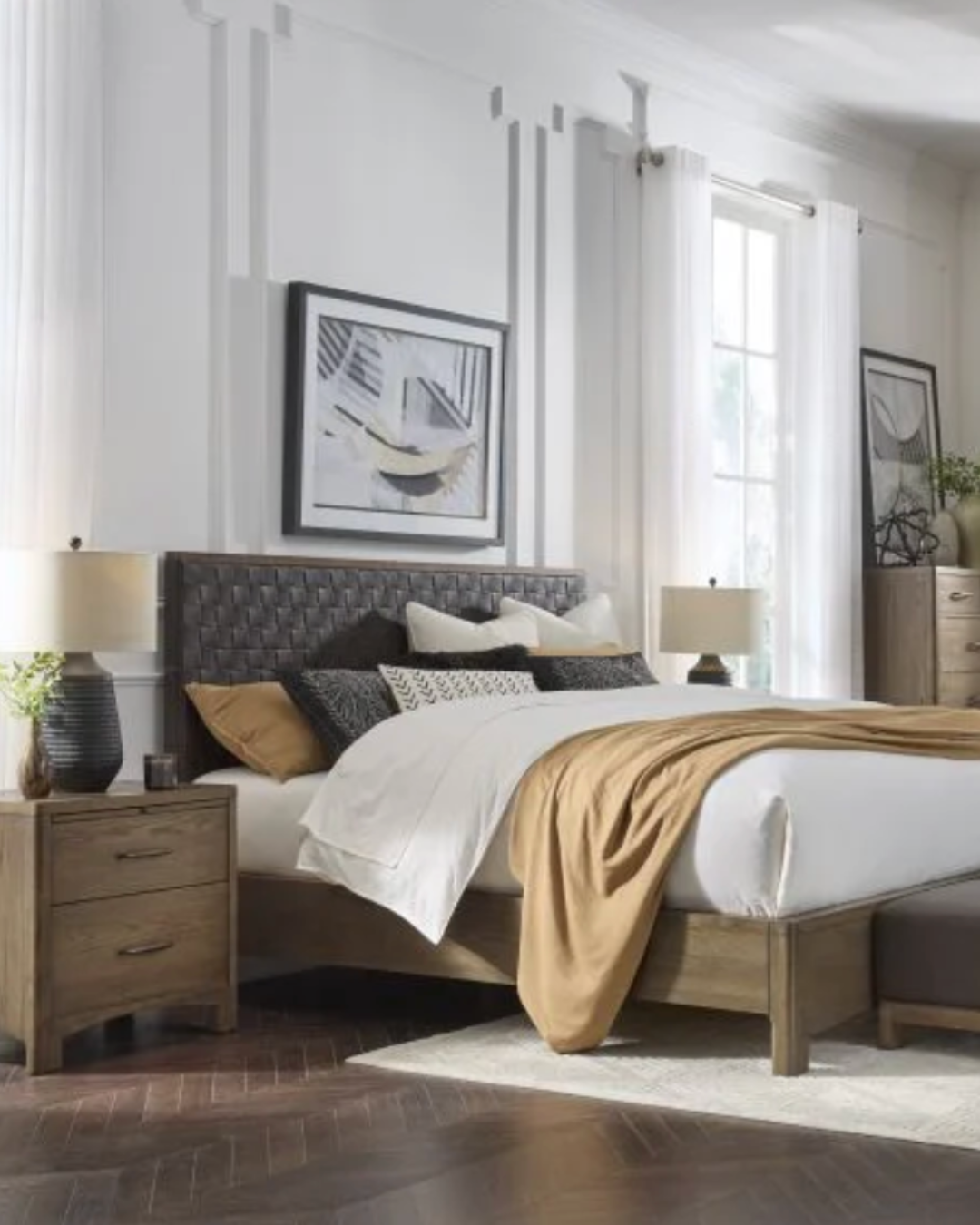 Habitación moderna con cama grande, cabecera tapizada en negro, dos mesitas con lámparas de noche, cortinas blancas, cuadros abstractos en la pared y muebles de madera clara.