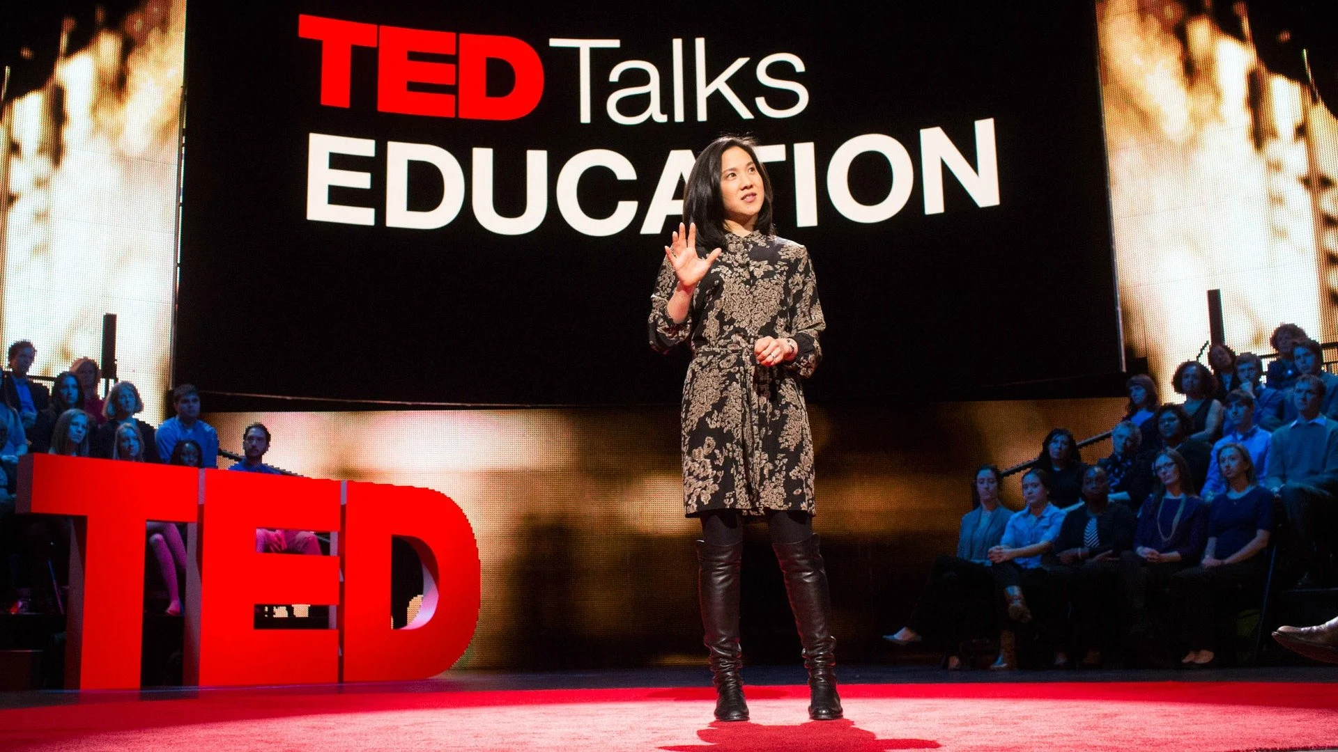 A woman giving a TED Talk on the stage with a large screen behind her displaying "TED Talks EDUCATION." The audience is seated and watching attentively.