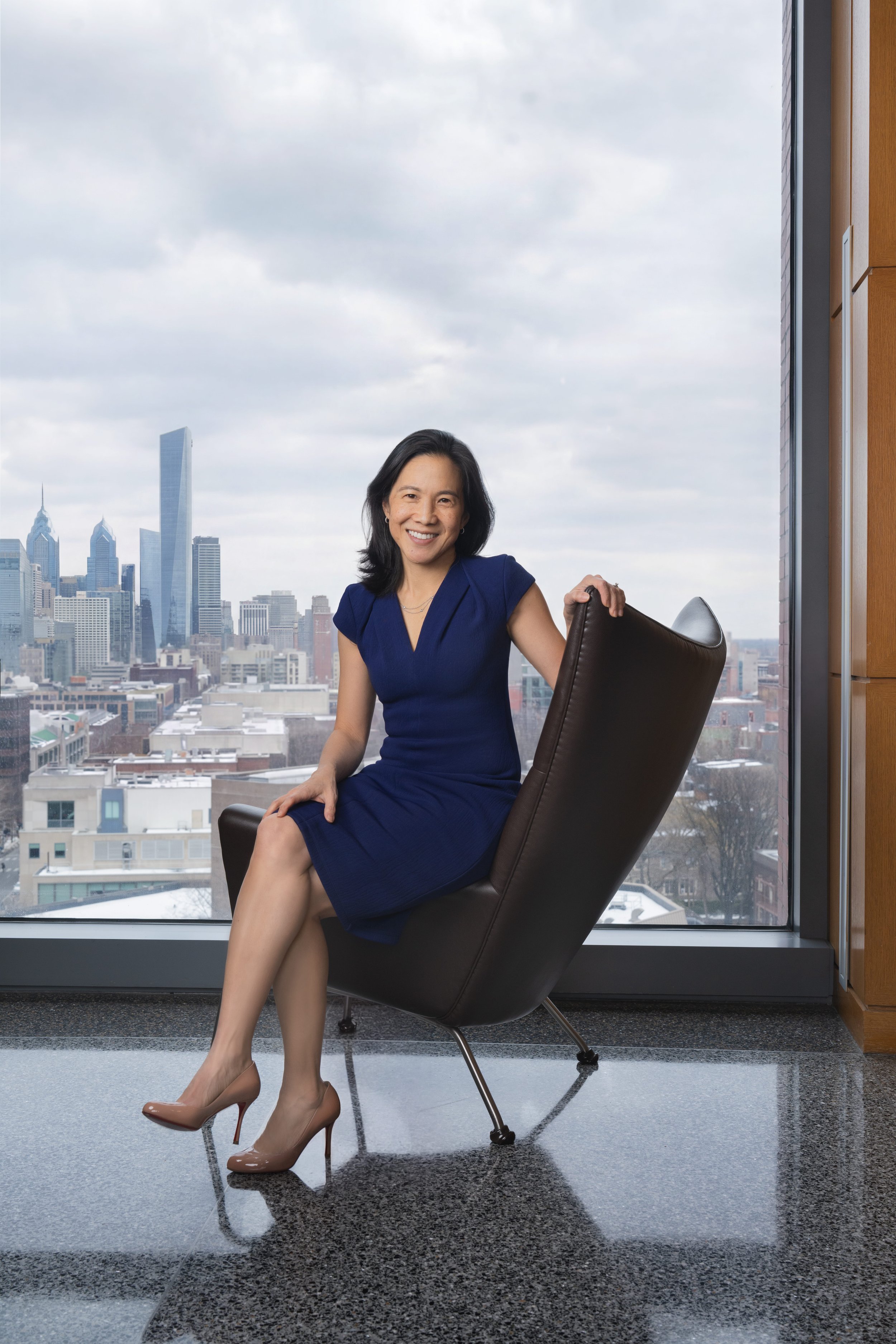 A woman with shoulder-length black hair, wearing a dark blue dress and nude high heels, sitting in a modern office chair near a large window with a city skyline view, smiling at the camera.