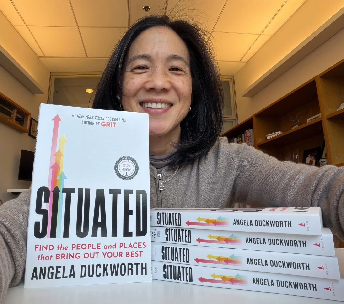 A woman takes a selfie with a book titled "Situated" by Angela Duckworth and multiple copies of the same book stacked in front of her. The book cover features colorful arrows and text highlighting key themes.