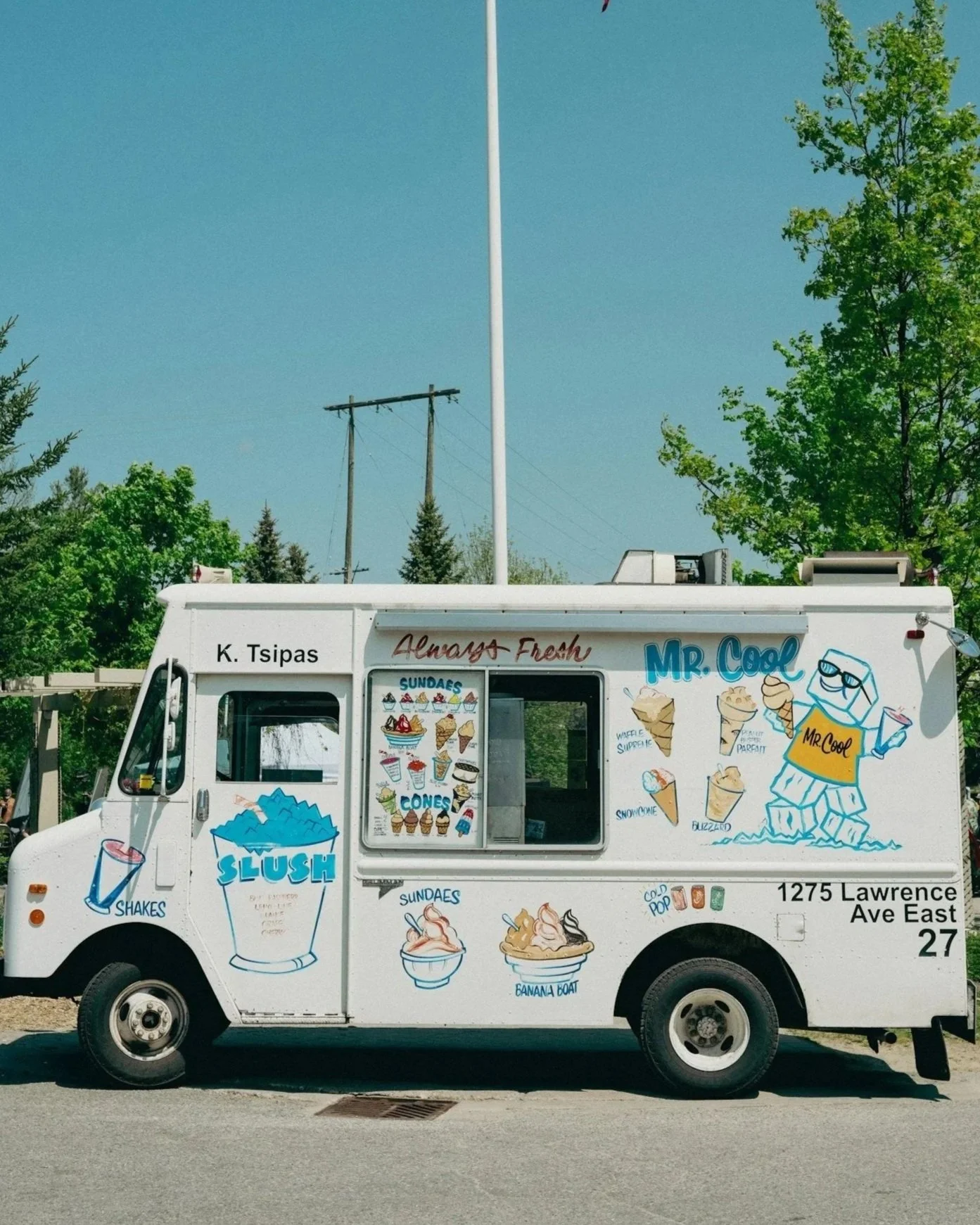 An ice cream truck advertising various frozen treats, including sundaes, cones, shakes, and slush drinks, parked on a street with trees and utility poles in the background.