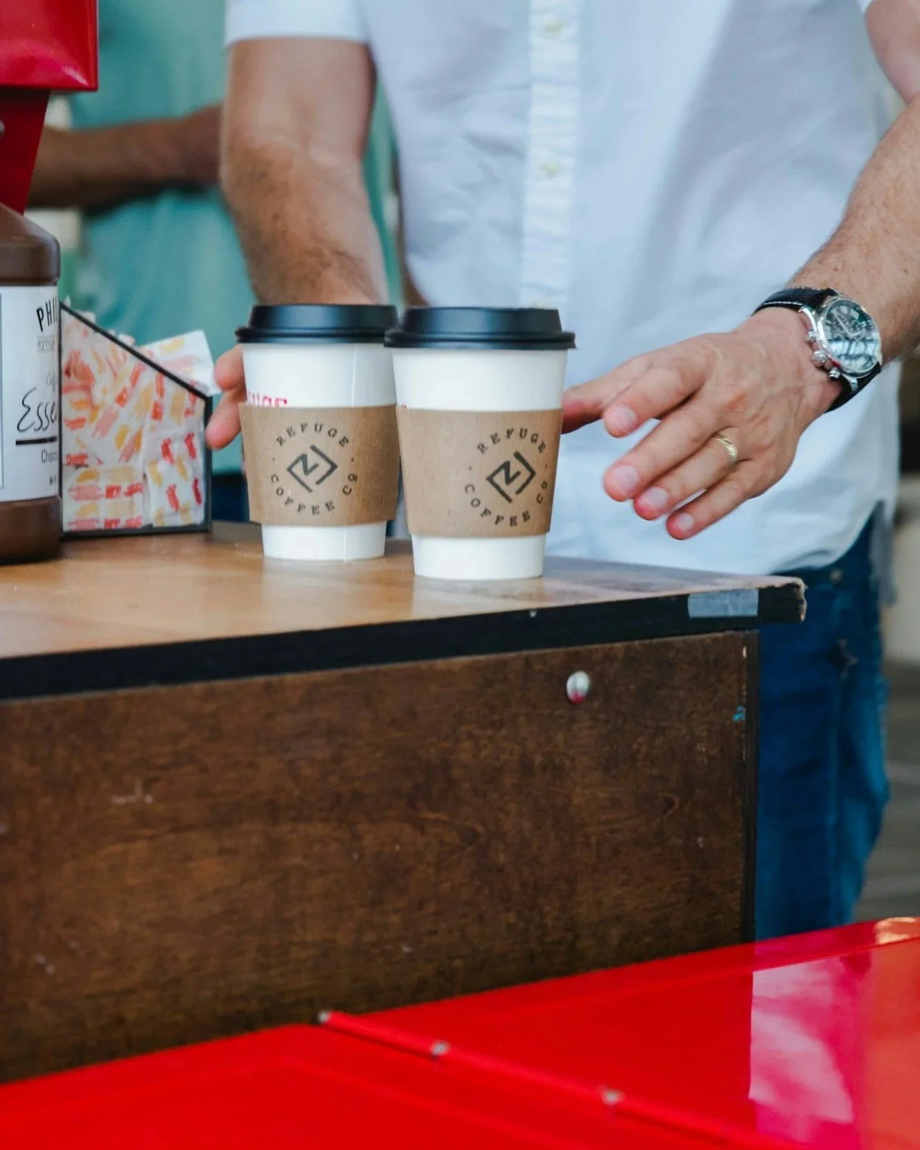 Two takeaway coffee cups with brown sleeves and black lids on a wooden counter, person in white shirt reaching for them, other items on the counter, indoors.