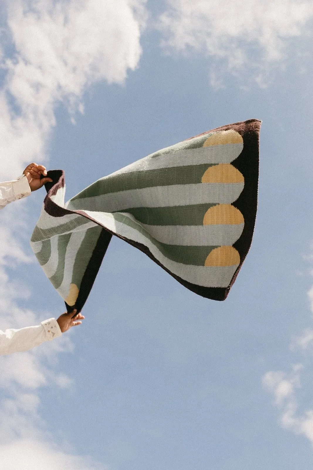 Two people holding up a colorful blanket with a sky and clouds background.