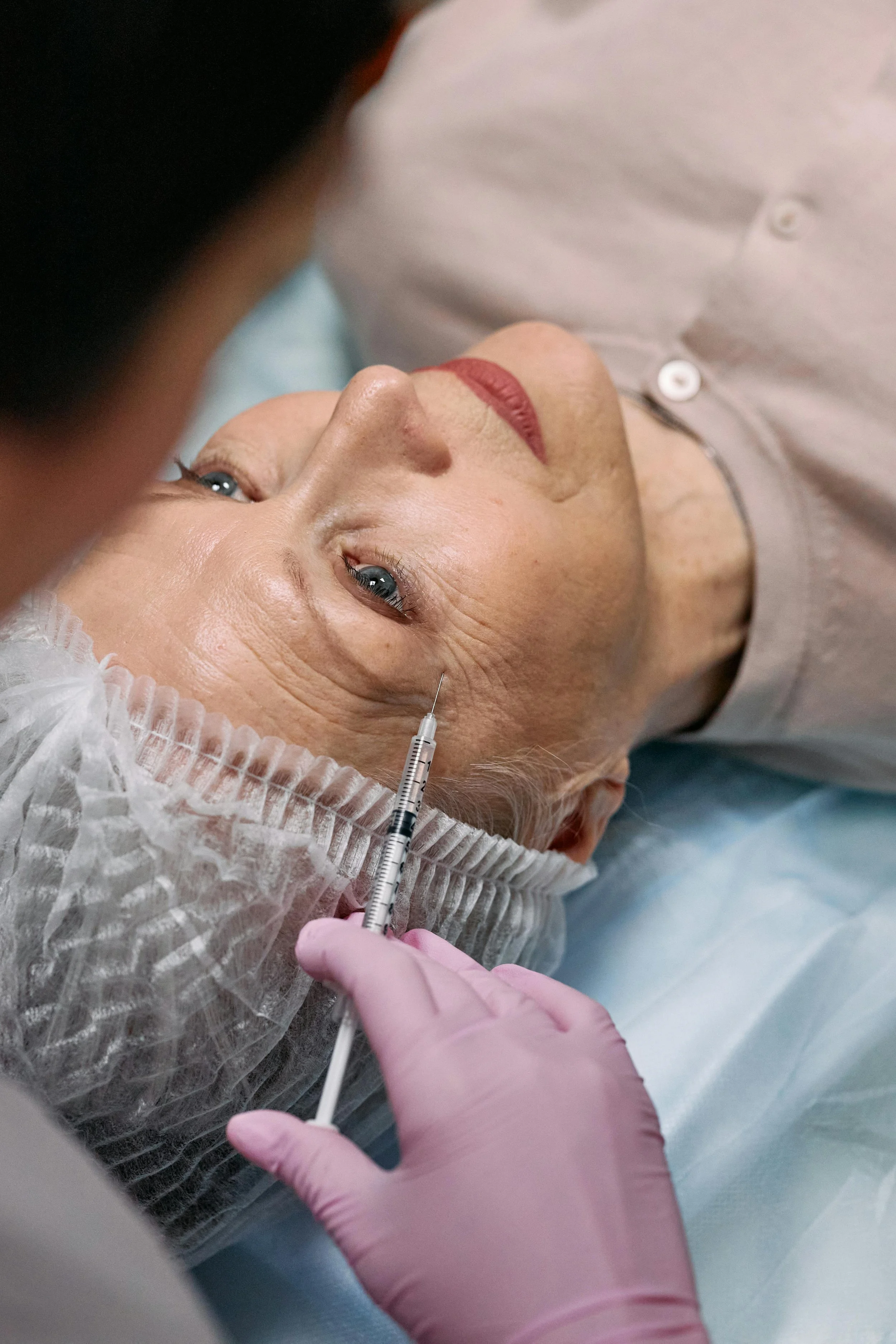 A woman lying on a medical bed receiving an injection in her face, with a healthcare professional wearing pink gloves administering the shot.